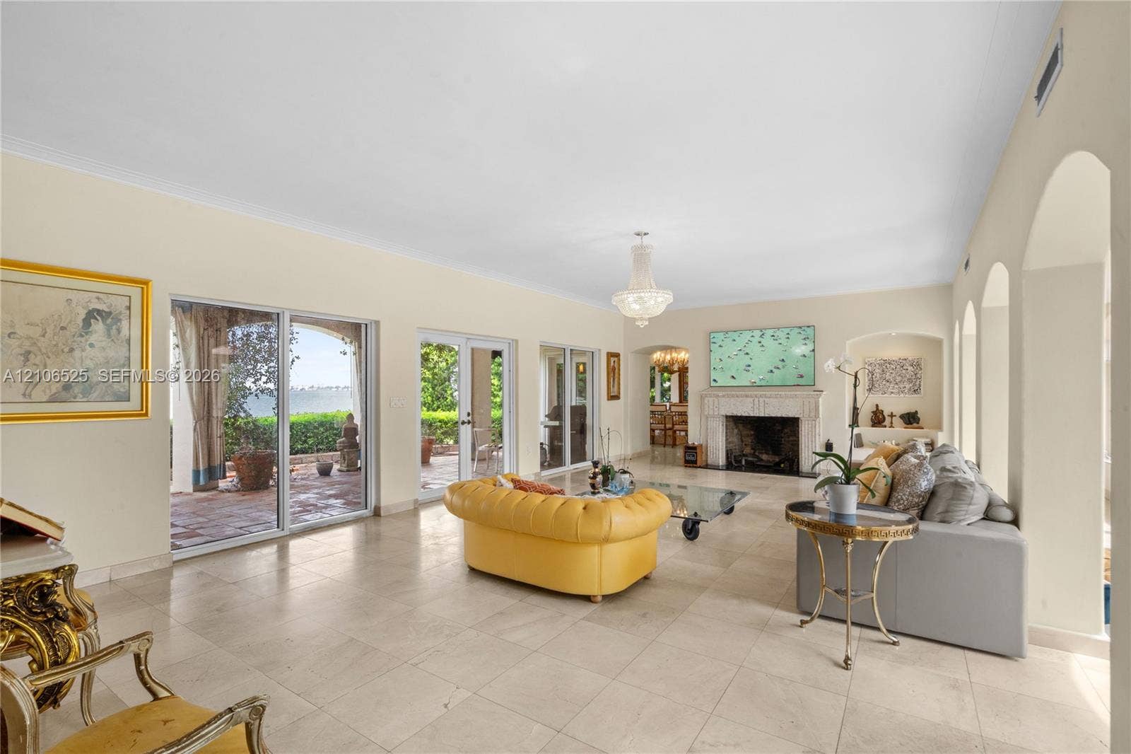 4760 N Bay Road, Miami Beach, FL 33140 Living area featuring large-format stone tile flooring, three sets of sliding glass doors, a stone-surround fireplace, arched alcoves, and a tiered crystal chandelier