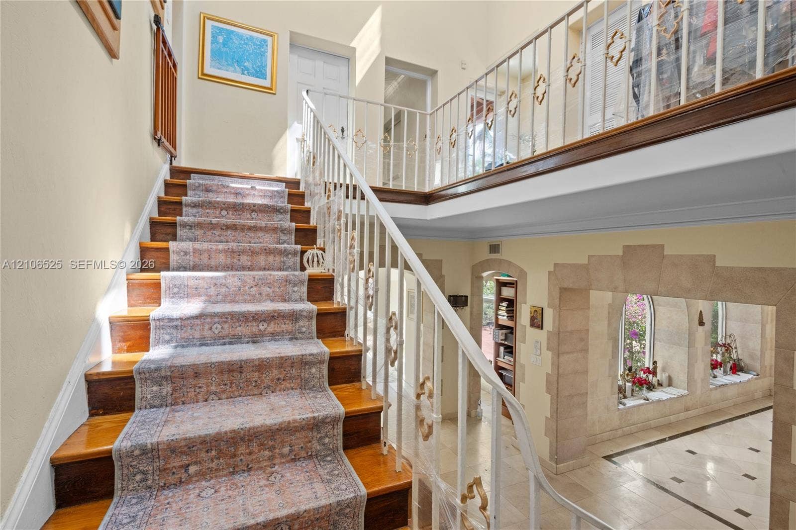 4760 N Bay Road, Miami Beach, FL 33140 Staircase featuring wood-finish treads, decorative metal railings, and a second-level balcony overlooking a stone-accented arched entryway with tiled flooring