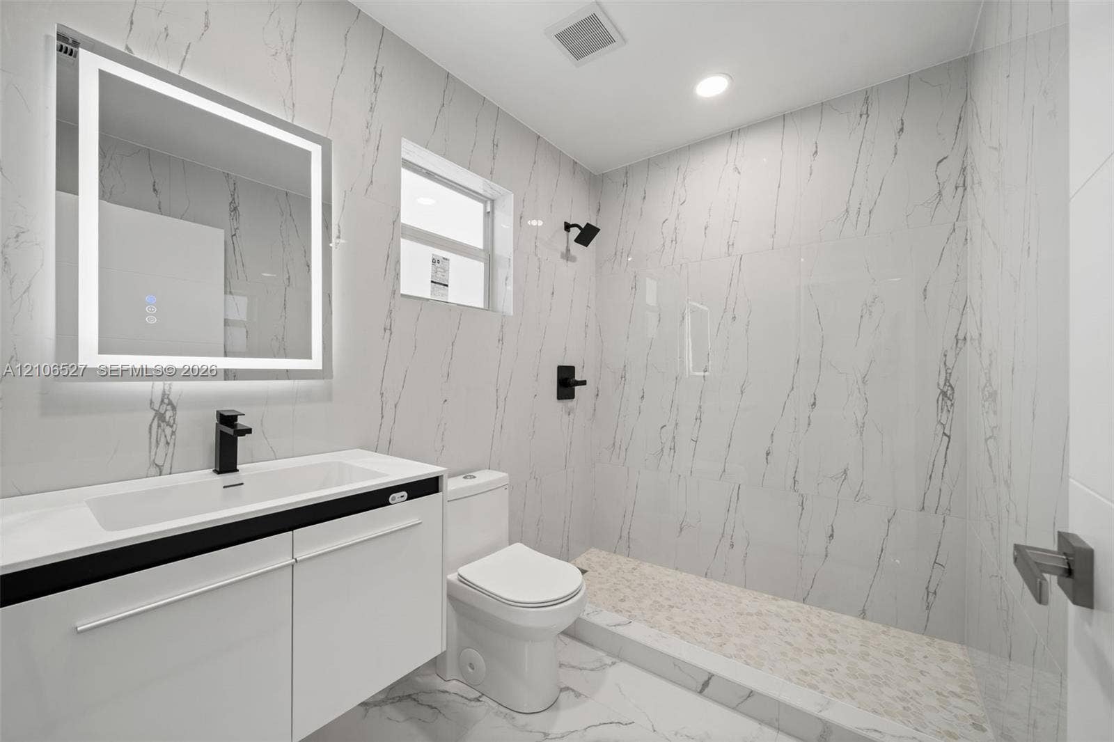 Bathroom featuring full-height stone-finish wall tile, walk-in shower with pebble-stone flooring, integrated LED vanity mirror, white cabinetry with a built-in sink, and matte black fixtures