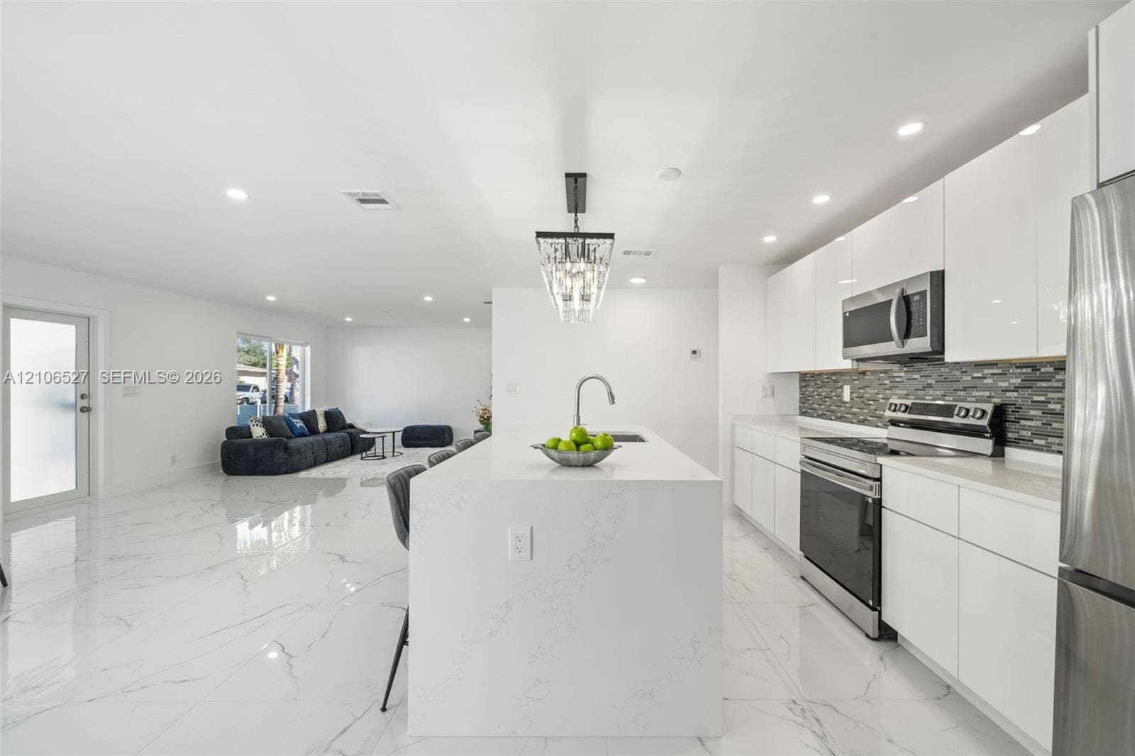 Kitchen featuring white flat-panel cabinetry, a central island with a solid-surface waterfall countertop, stainless steel appliances, a glass-tile backsplash, and polished stone-finish flooring