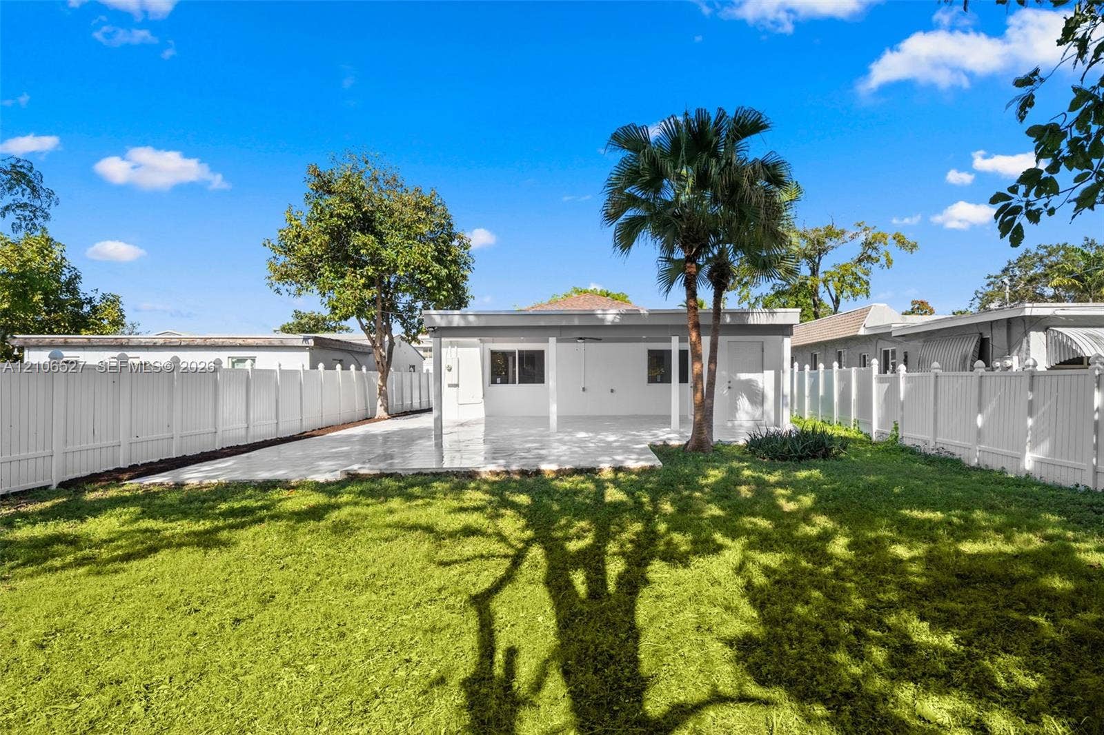 Rear exterior featuring a covered patio, paved hardscaping, white vinyl privacy fencing, and mature palm trees
