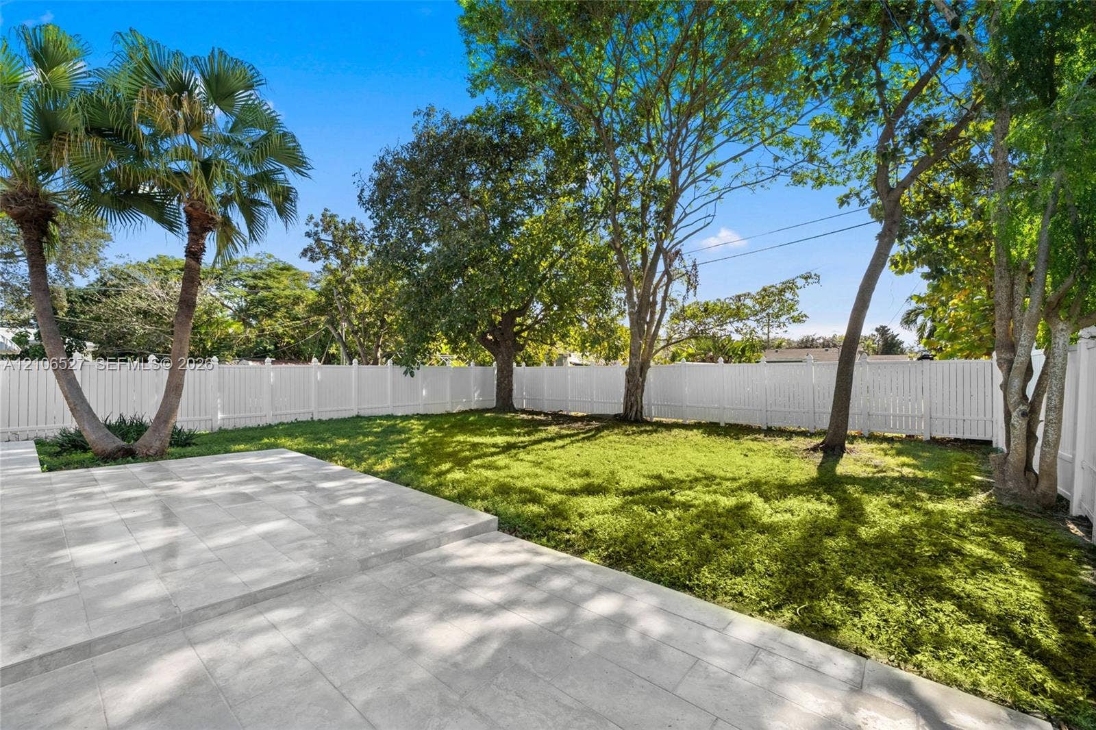 Backyard featuring a paved stone patio, mature trees, manicured lawn, and a full-perimeter white vinyl fence