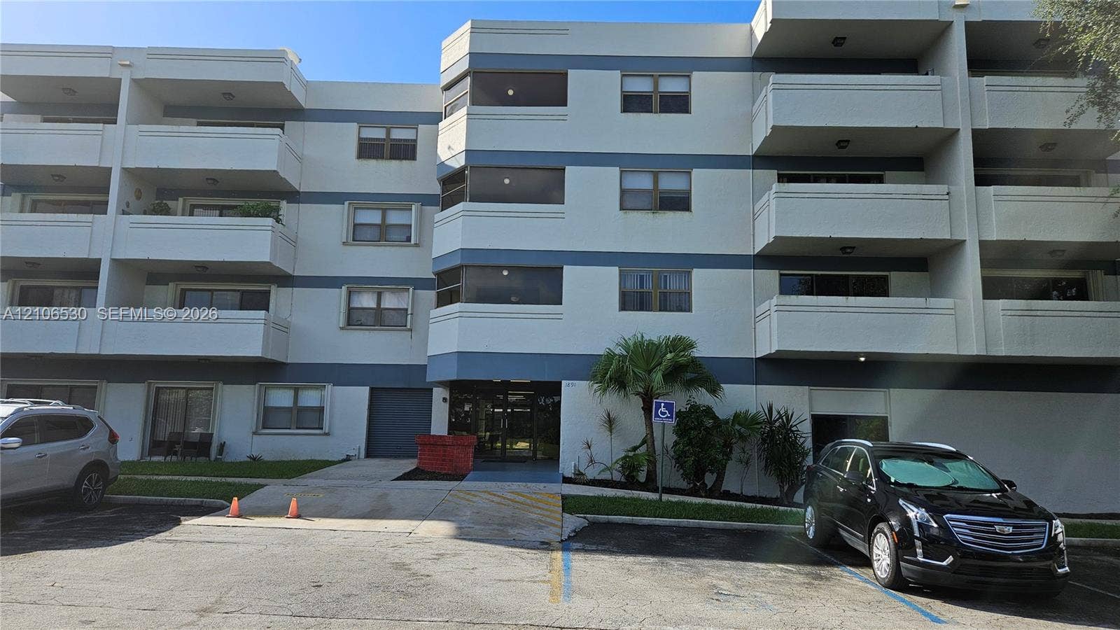 Multi-story residential building exterior featuring a concrete facade with horizontal detailing, individual private balconies, glass-pane windows, and paved surface parking