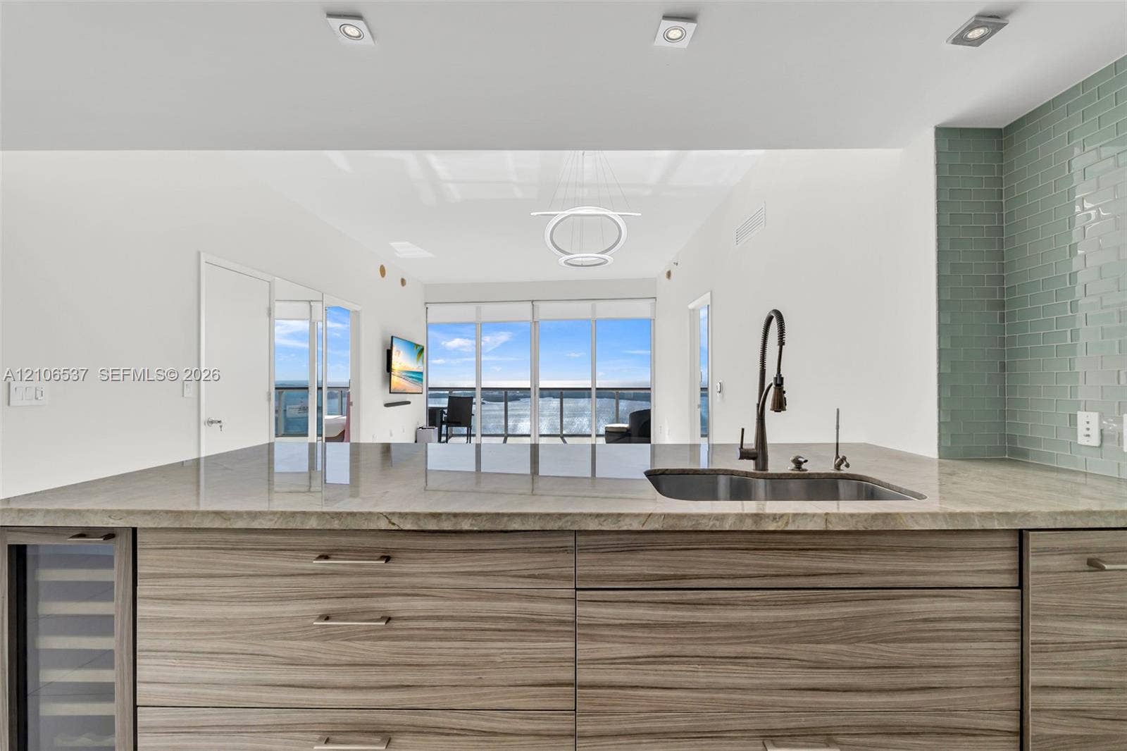 465 Brickell Avenue, #5203, Miami, FL 33131 Kitchen island featuring wood-finish cabinetry, stone countertop, undermount sink with a matte black pull-down faucet, and a built-in wine cooler, complemented by a light green subway tile backsplash