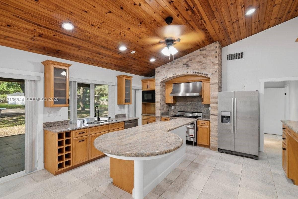 Vaulted wood-plank ceiling featuring recessed lighting and a central ceiling fan, stone-accented kitchen range surround, stone-top kitchen island, light-toned tile flooring, and stainless steel kitchen appliances