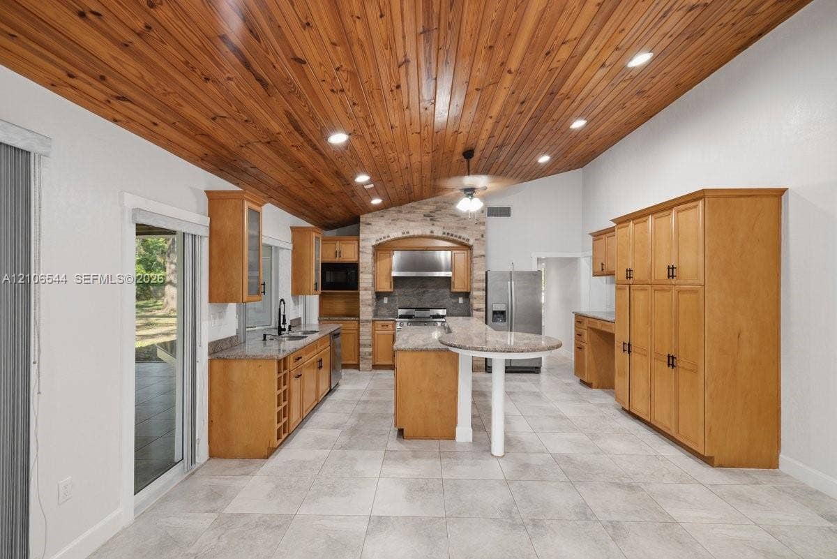 Vaulted wood-plank ceiling featuring recessed lighting and a central ceiling fan, stone-accented kitchen range surround, stone-top kitchen island, light-toned tile flooring, and stainless steel kitchen appliances
