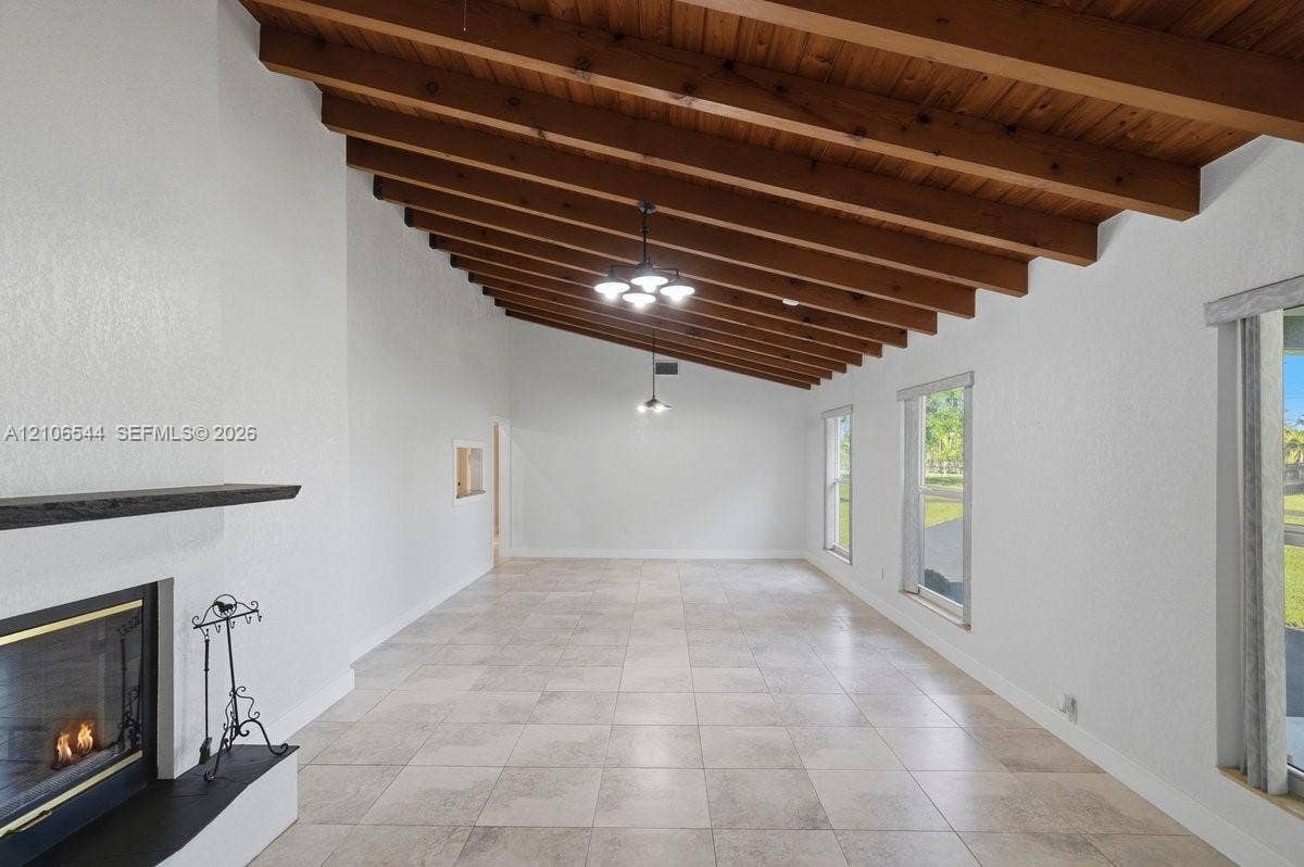 Vaulted ceiling with exposed wood beams, light-colored tile flooring, stone-surround fireplace, and multi-light pendant fixtures
