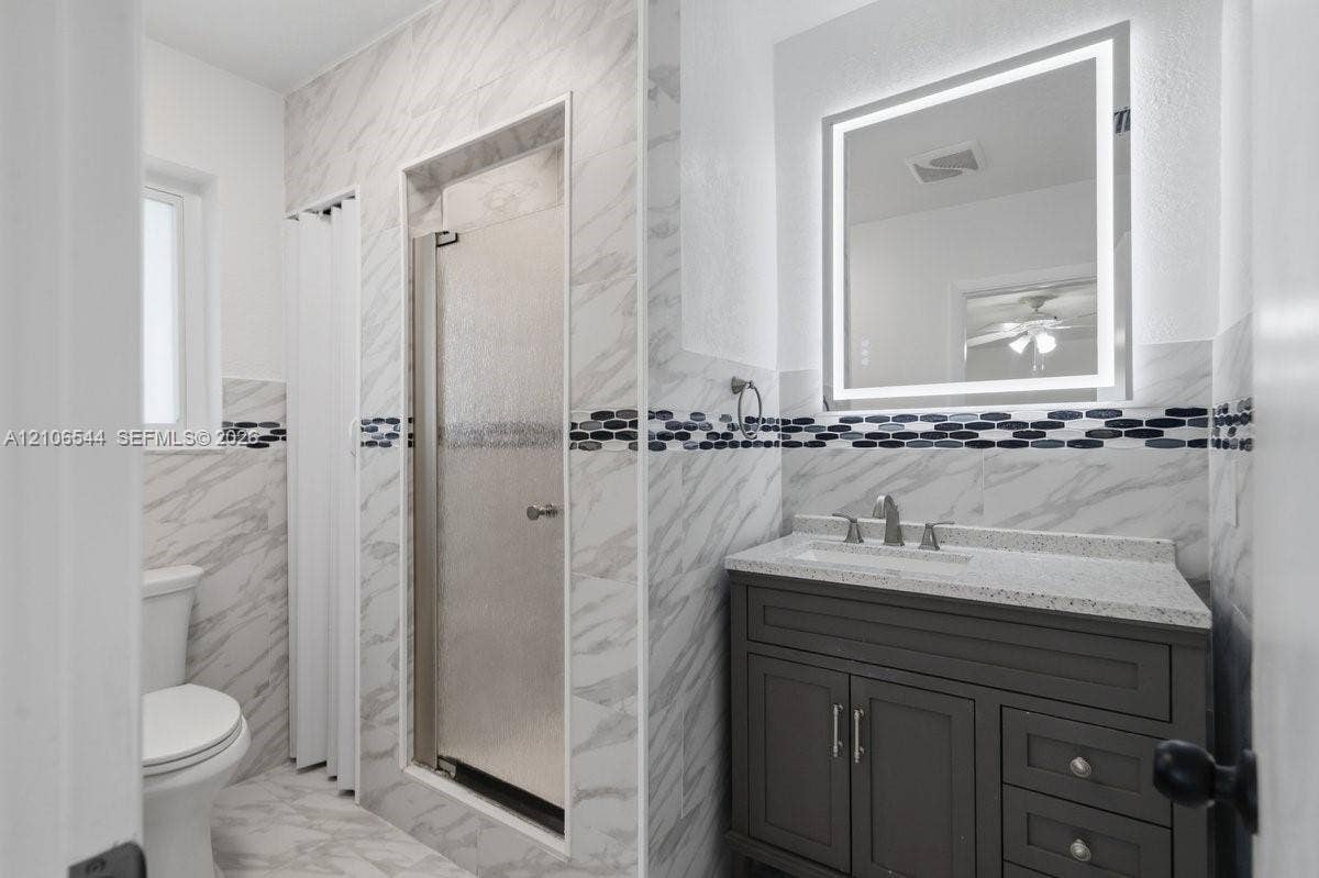 Bathroom featuring stone-finish wall tile with horizontal decorative accent inlay, enclosed shower with textured glass door, dark cabinetry with light-toned countertop, and integrated-light vanity mirror