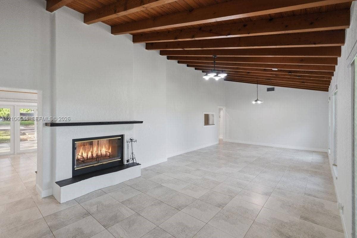 Vaulted ceiling with exposed wood beams, neutral tile flooring, fireplace with a black mantel, and two hanging pendant light fixtures