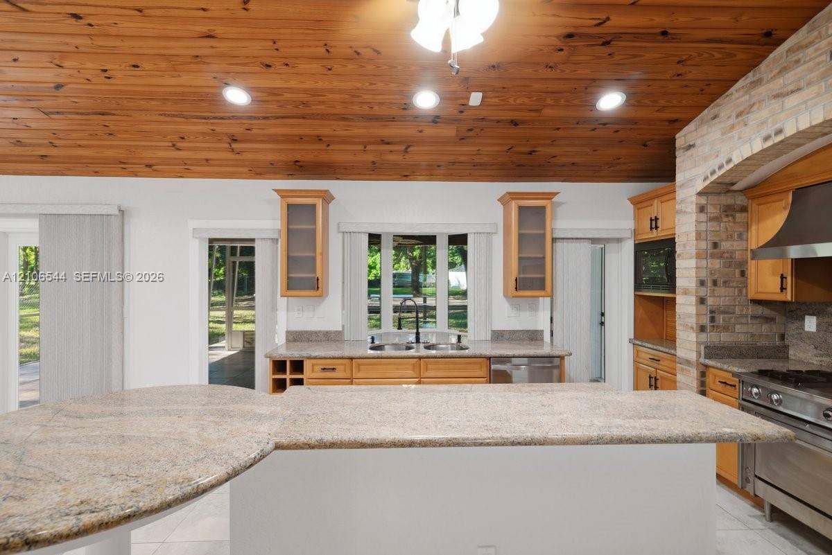 Kitchen featuring vaulted wood-plank ceiling, natural stone countertops, custom brick archway, stainless steel range with matching hood, and warm-toned cabinetry