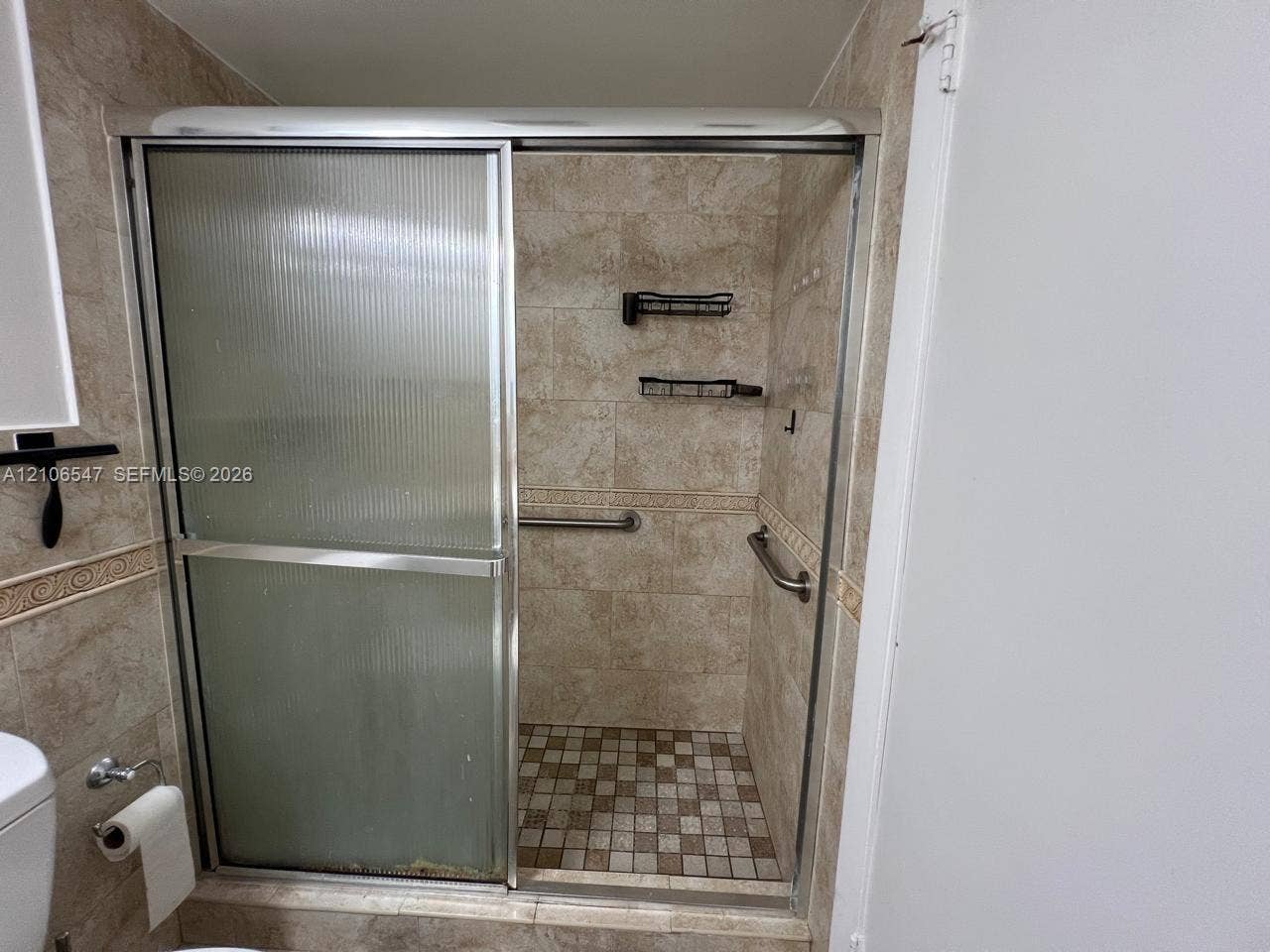 6801 Indian Creek Drive, #509, Miami Beach, FL 33141 Walk-in shower enclosure featuring ribbed sliding glass doors, neutral-toned tiled walls with decorative border accents, mosaic tile flooring, and integrated metal grab bars