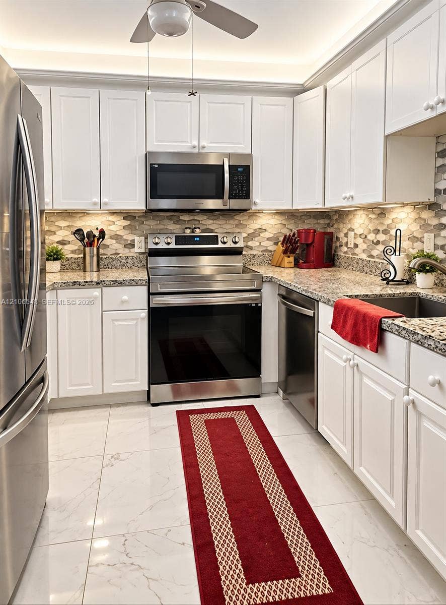 Kitchen featuring white shaker-style cabinetry, stone-finish countertops, stainless steel appliances, a hexagonal tile backsplash, and large-format tile flooring