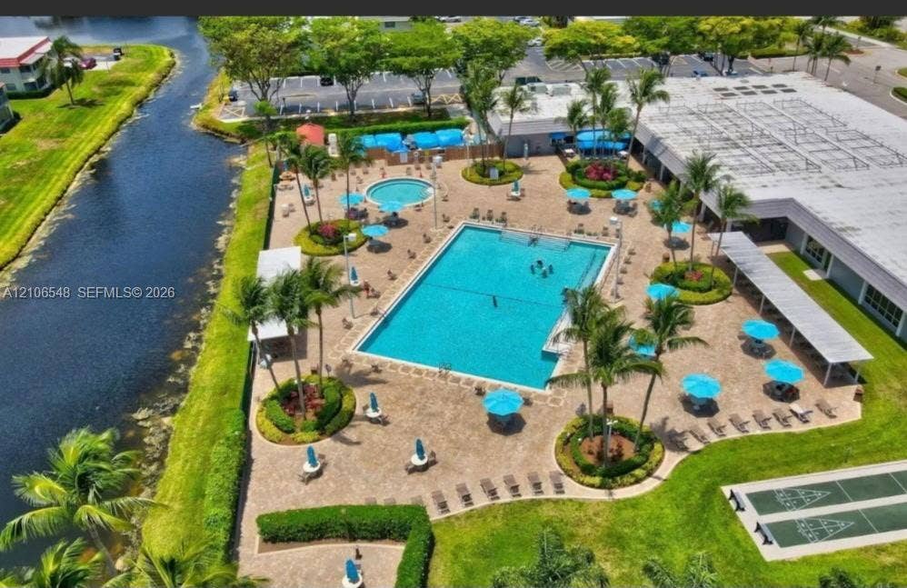 Outdoor recreation area featuring a rectangular swimming pool, separate circular spa, paved pool deck, and landscaped grounds with mature palm trees and shuffleboard courts