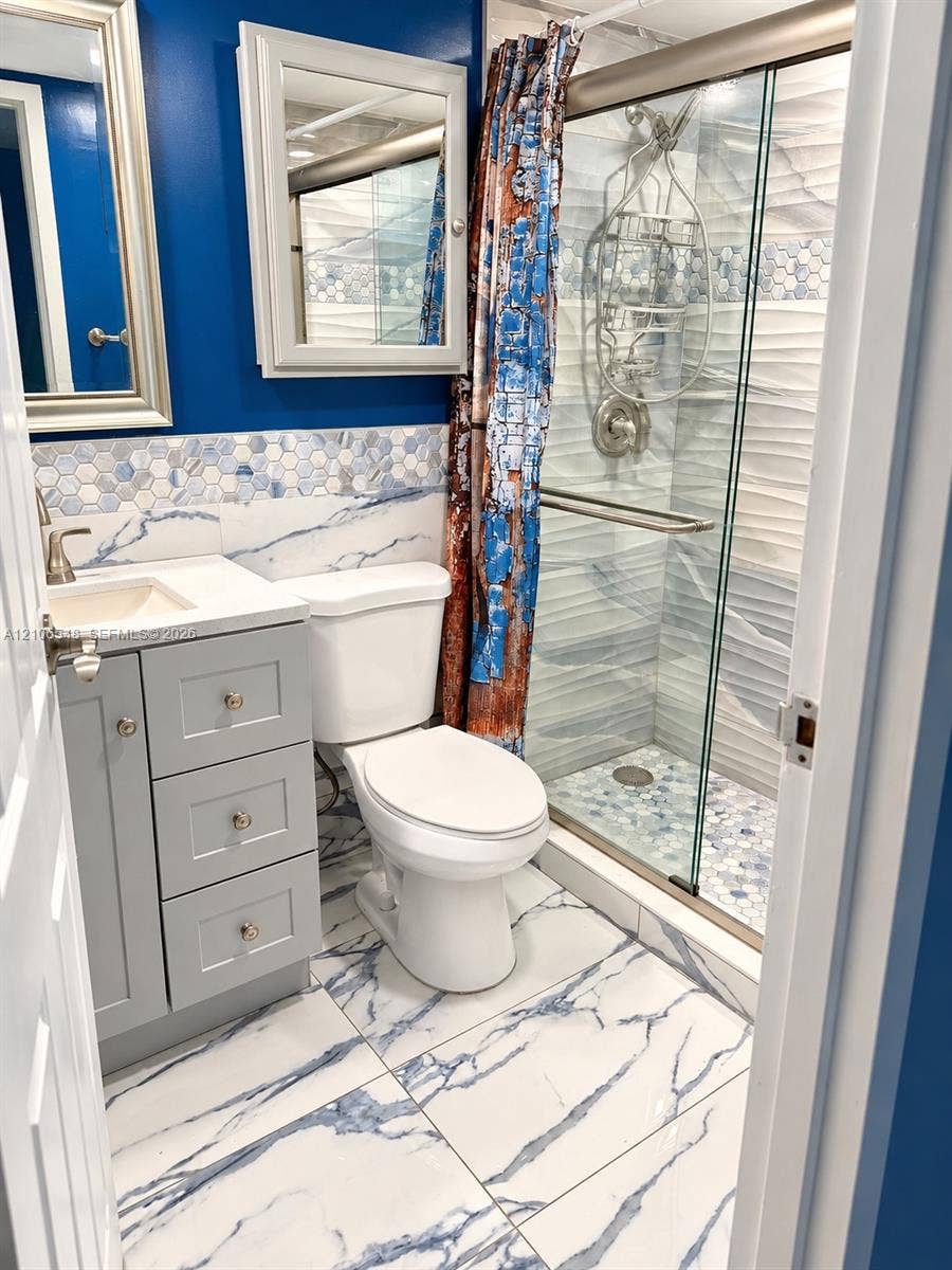 Grey vanity with white countertop, shower enclosure with glass sliding doors, stone-finish floor tiling, patterned wall tile accents, and wall-mounted medicine cabinet