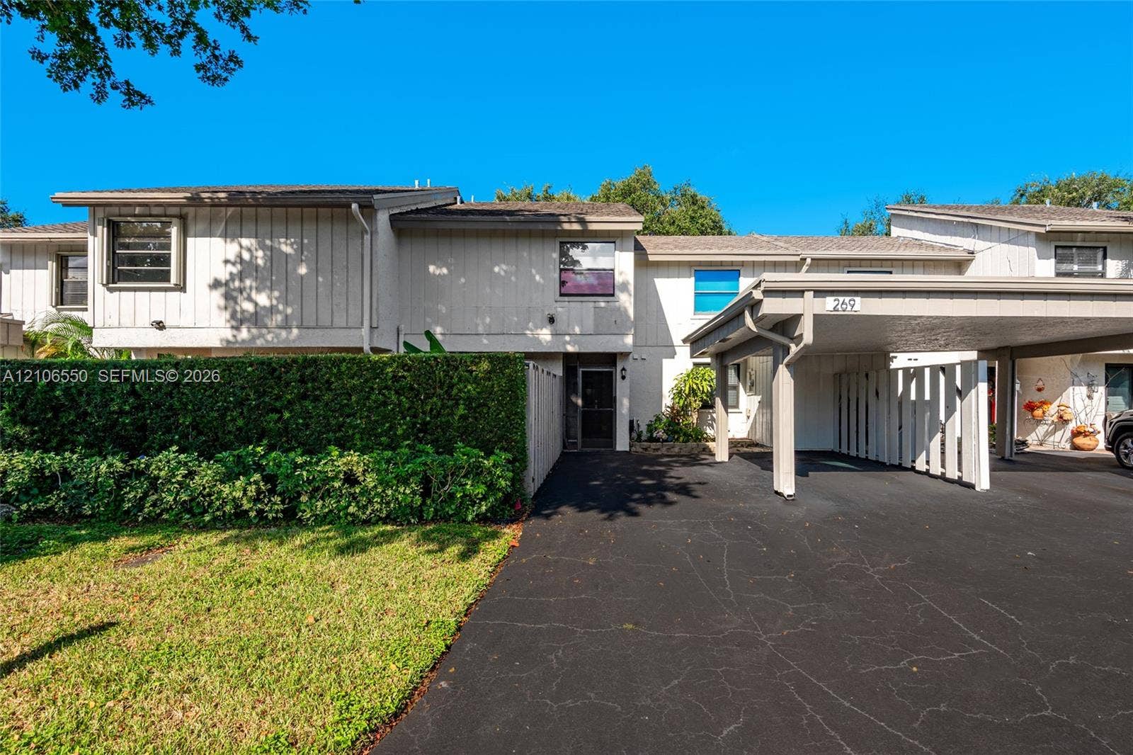 Exterior facade featuring vertical siding, a paved driveway, attached covered carport, and a manicured privacy hedge