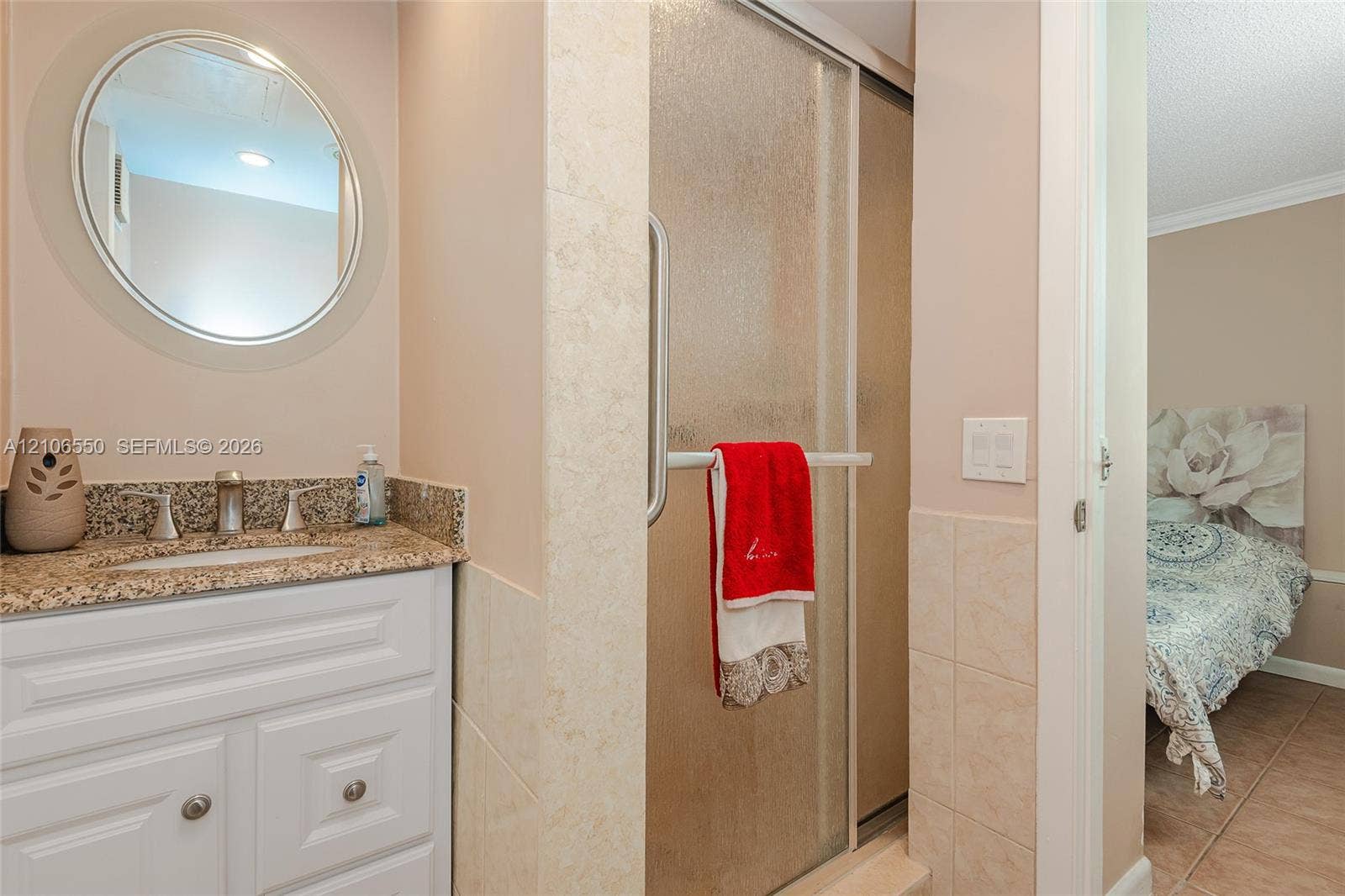 White cabinetry vanity with stone-finish countertop, undermount sink, circular wall-mounted mirror, and glass-enclosed shower with tiled surround