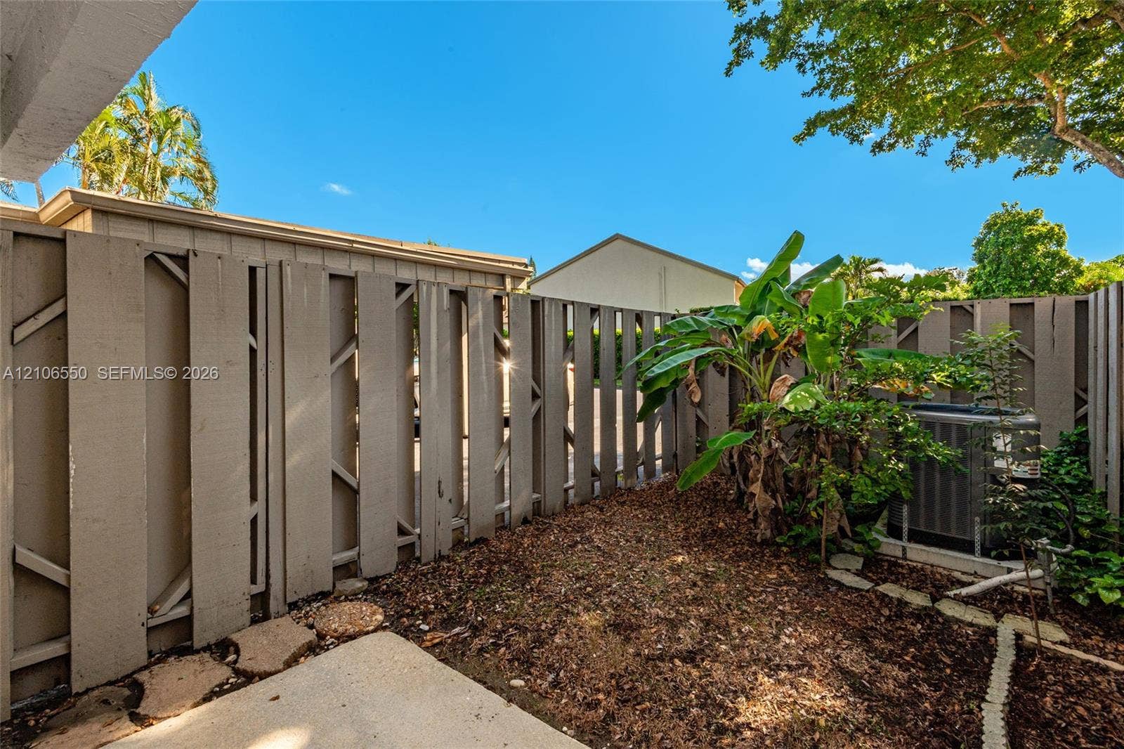 Private outdoor space featuring vertical wood-slat privacy fencing, concrete patio slab, mulch ground cover, and mature tropical greenery