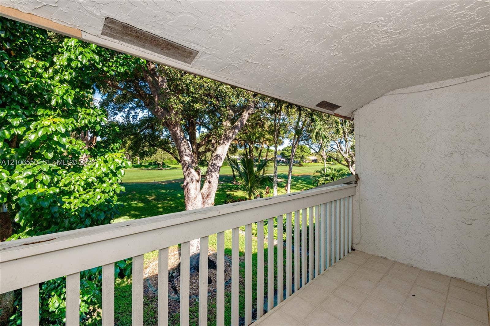 Covered balcony featuring tile flooring, textured stucco wall, and wooden balustrade railing, overlooking mature trees and landscaped grounds
