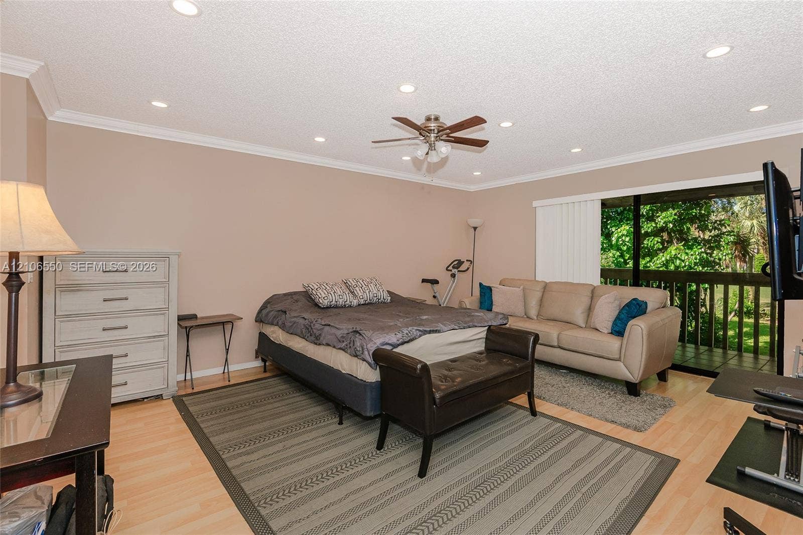 Light-toned wood-finish flooring, recessed ceiling lighting, central ceiling fan with integrated light fixture, crown molding, and sliding glass doors with access to a railed balcony