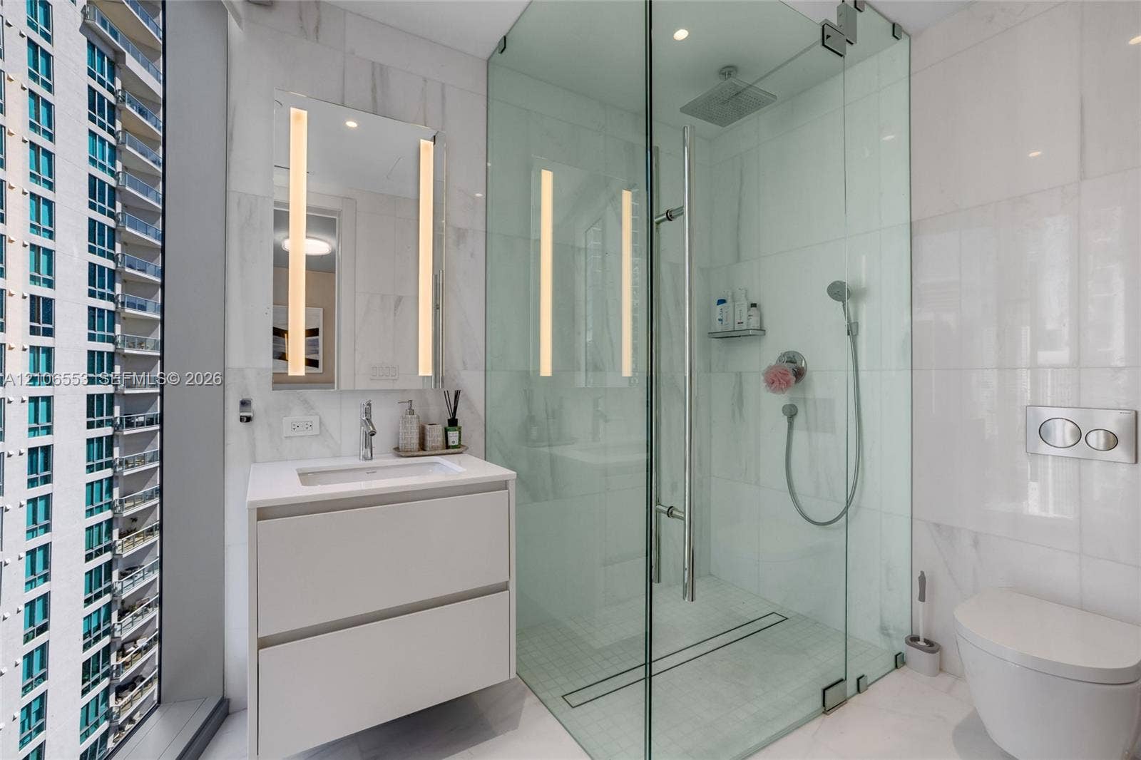 300 Biscayne Boulevard Way, #3002E, Miami, FL 33131 Bathroom featuring a floating vanity with a rectangular sink, wall-mounted mirror with integrated lighting, glass-enclosed shower with rainfall head and handheld sprayer, wall-mounted toilet, and floor-to-ceiling tile finishes