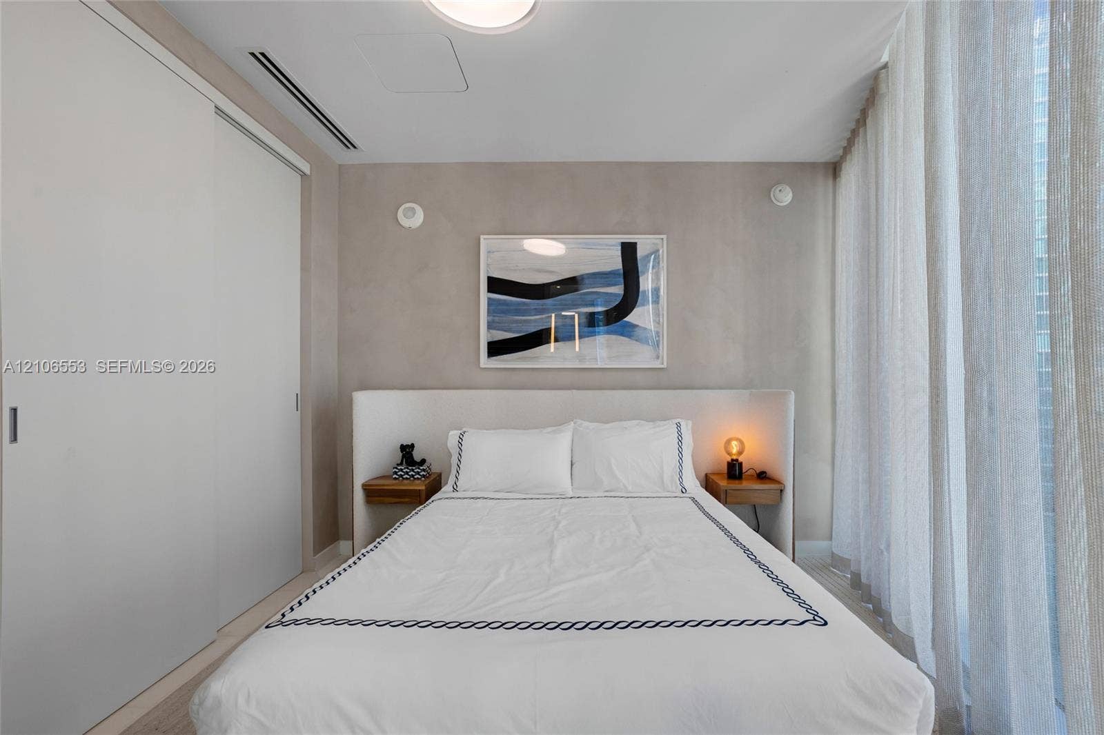 300 Biscayne Boulevard Way, #3002E, Miami, FL 33131 Neutral-toned room featuring a built-in upholstered headboard, dual integrated wood-finish bedside shelves, floor-to-ceiling sliding closet doors, and light-filtering floor-to-ceiling drapery