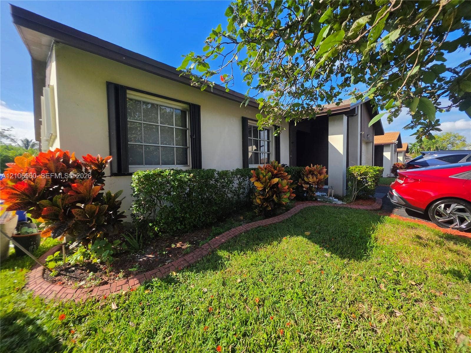 9550 W Elm Lane, Miramar, FL 33025 Stucco exterior featuring grid-pattern windows, a brick-edged garden bed, mature landscaping, and an asphalt driveway
