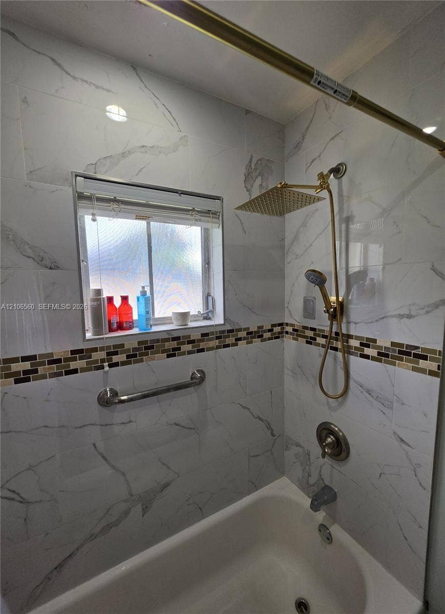 9550 W Elm Lane, Miramar, FL 33025 Bathtub surround featuring floor-to-ceiling stone-pattern tile, a multi-tone mosaic accent border, a gold-tone rainfall showerhead with a handheld sprayer, and a mounted metal grab bar