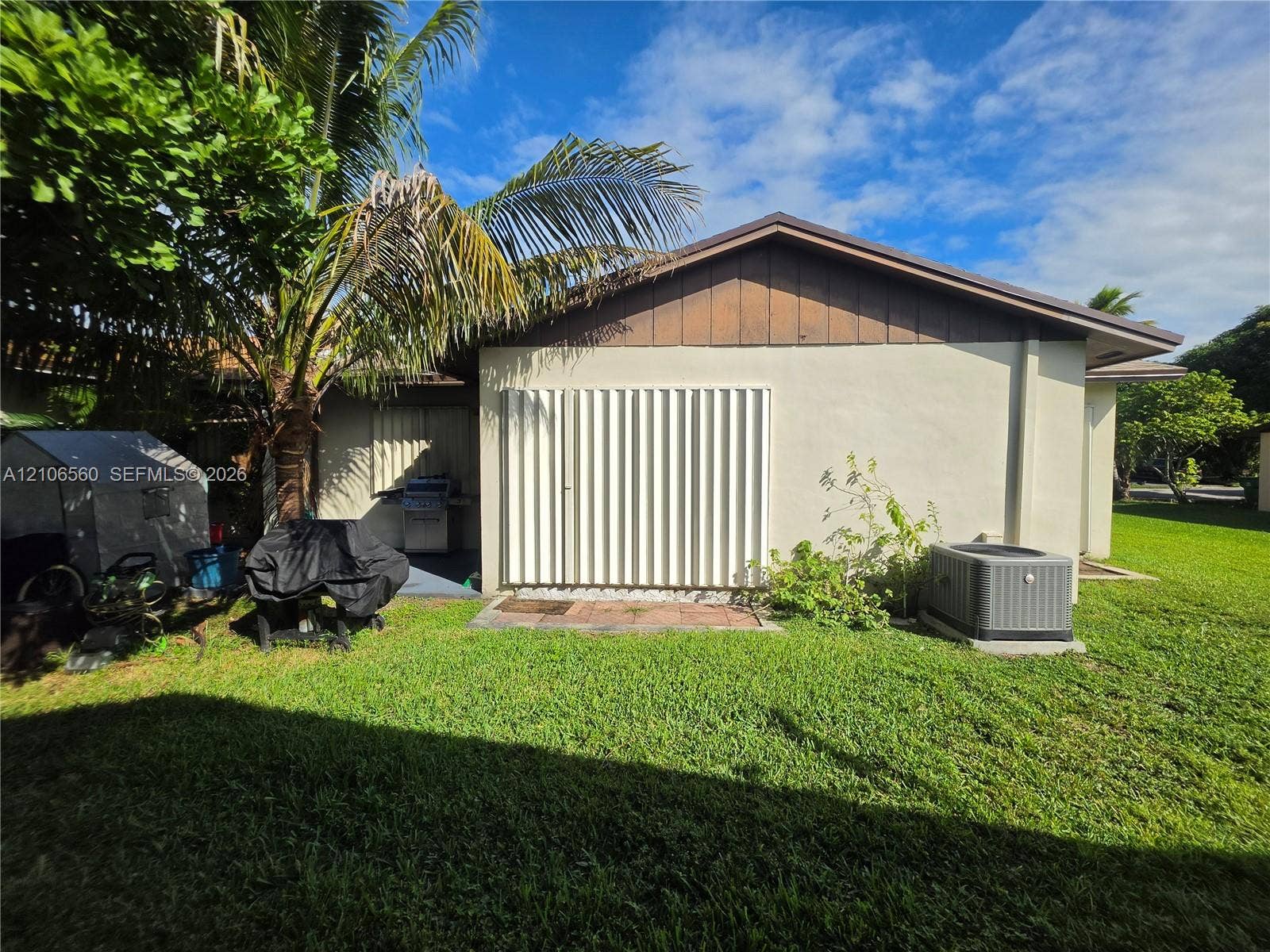 9550 W Elm Lane, Miramar, FL 33025 Exterior side profile featuring light-toned stucco walls, vertical wood siding, integrated accordion-style shutters, and a rear lawn