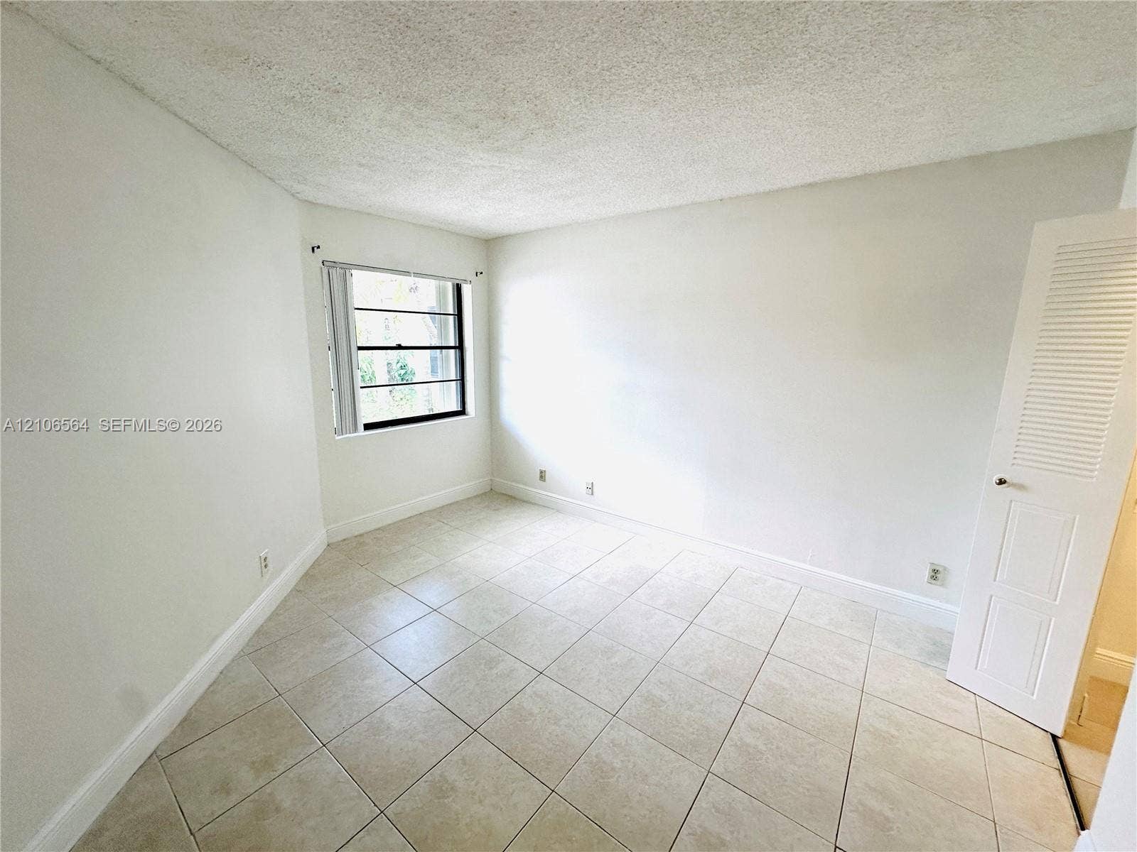 Room featuring neutral tile flooring, light-toned wall finishes, a window with horizontal blinds, and a louvered closet door