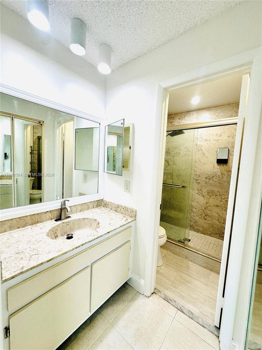 Vanity featuring a stone-composite countertop with an integrated basin, large wall-mounted mirror, overhead cylindrical track lighting, and a glass-enclosed shower with tile surround