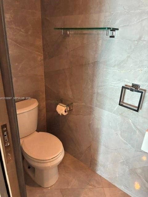 Bathroom featuring large-format tile wall cladding, a porcelain toilet, a glass wall-mounted shelf, and chrome hardware