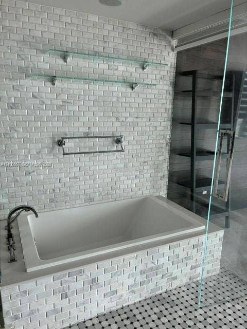 Built-in soaking tub with a tiled surround, complemented by a floor-to-ceiling accent wall in brick-pattern stone, dual glass wall-mounted shelves, and a wall-mounted metal towel rack