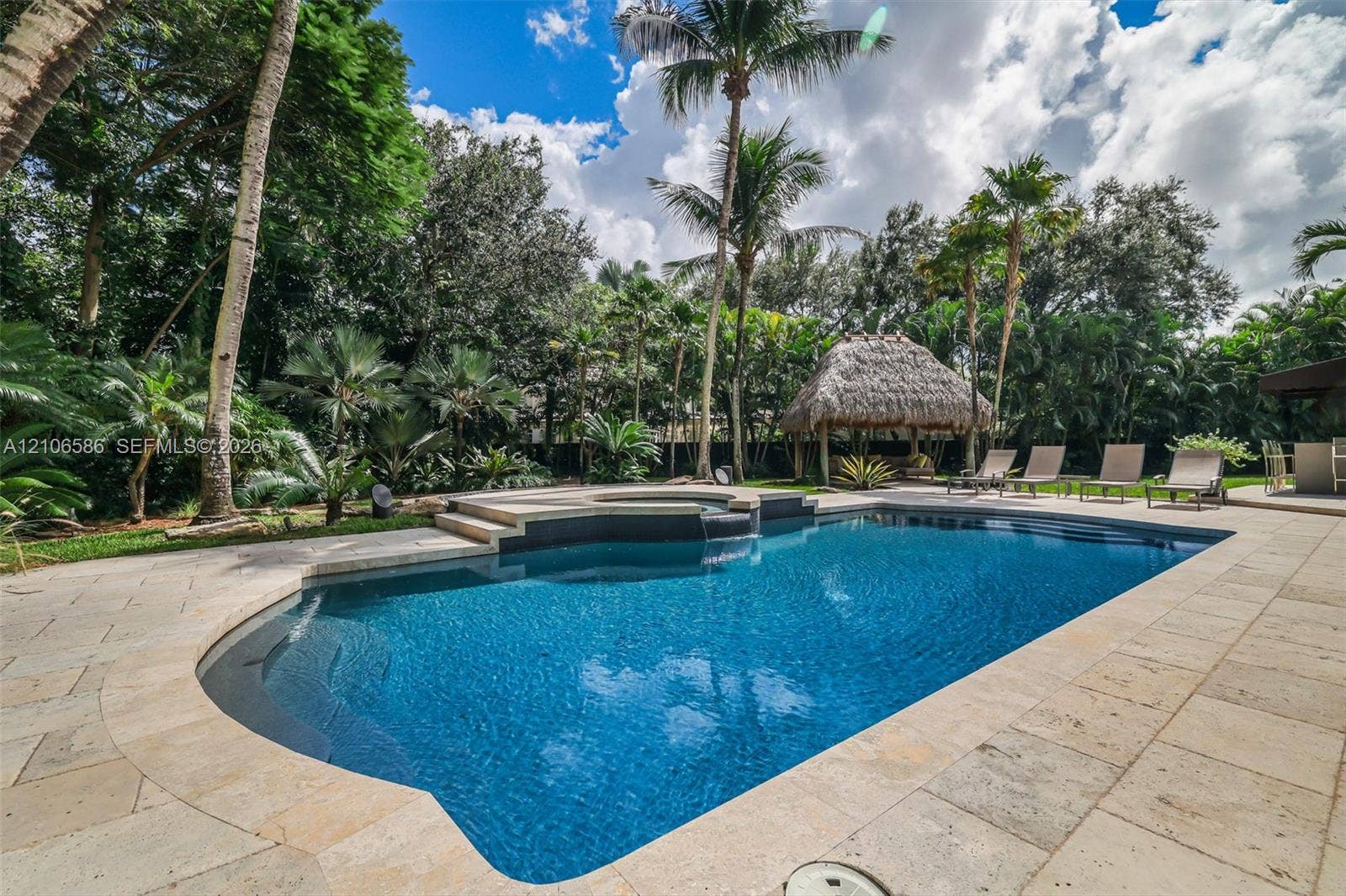 9325 SW 59th Place, Pinecrest, FL 33156 In-ground swimming pool with an integrated elevated spa, stone paver decking, and a detached thatched-roof structure surrounded by mature palms and landscaping