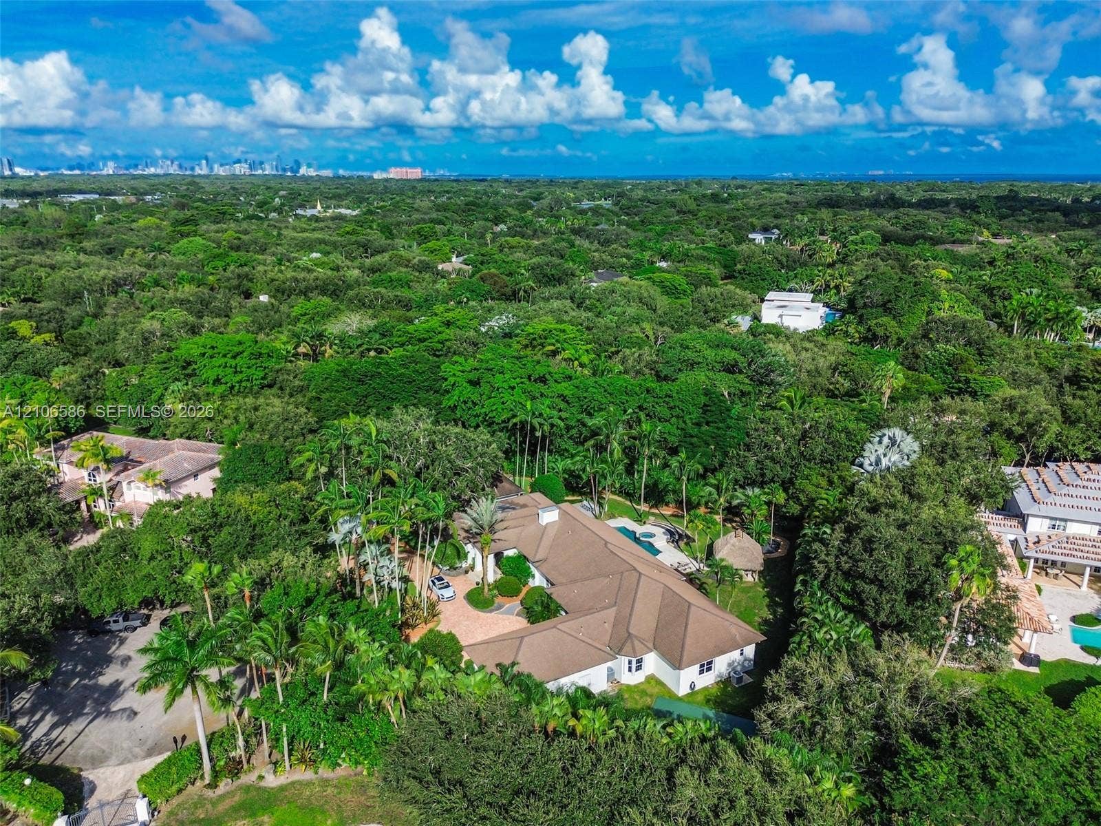 9325 SW 59th Place, Pinecrest, FL 33156 Aerial perspective featuring a multi-gabled roofline, a circular paved driveway, a private swimming pool, and extensive mature landscaping on a heavily wooded lot