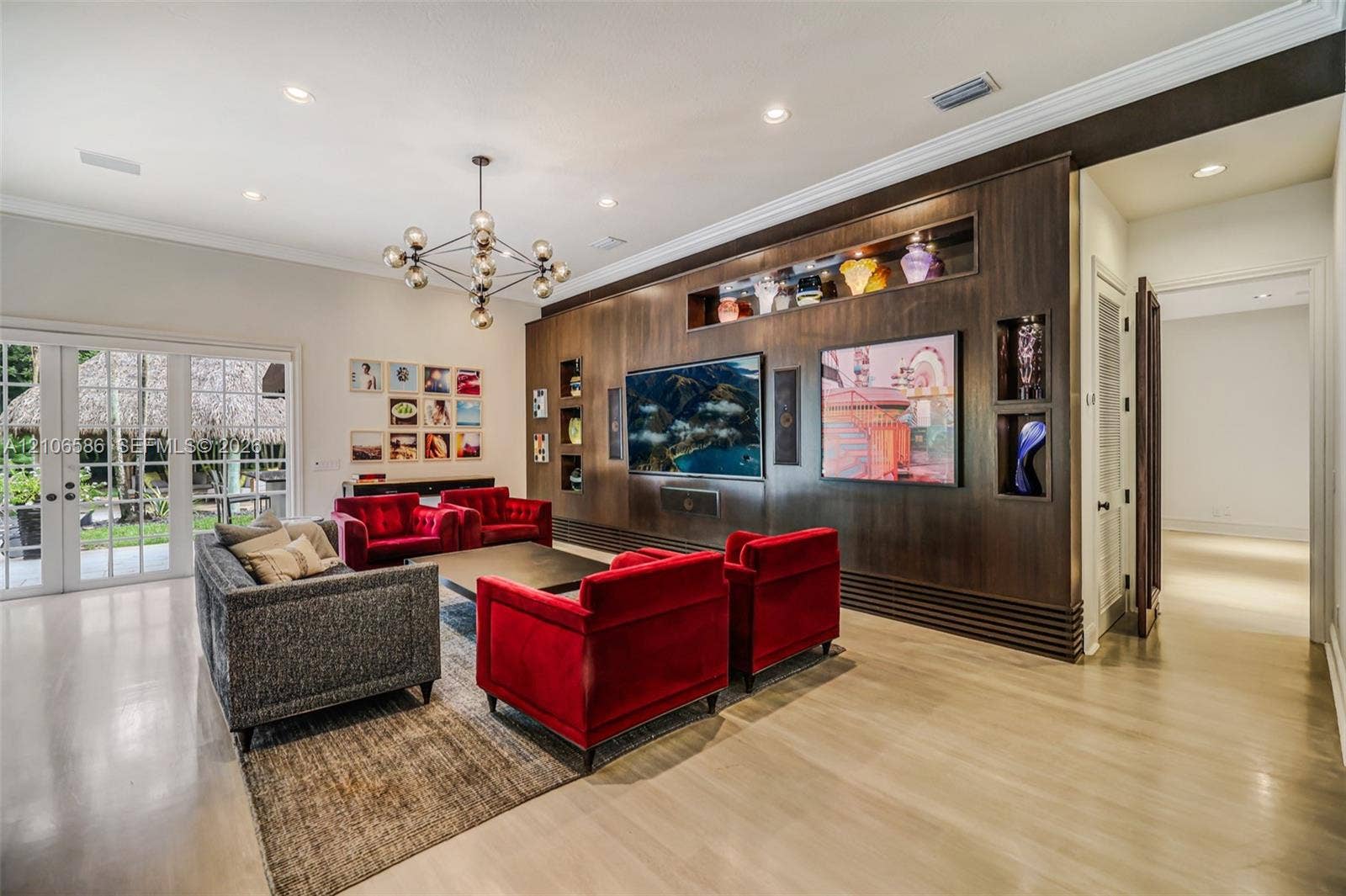 9325 SW 59th Place, Pinecrest, FL 33156 Custom wood-veneer accent wall featuring integrated recessed shelving, multi-arm geometric chandelier, light-toned flooring, and floor-to-ceiling glass-paneled double doors
