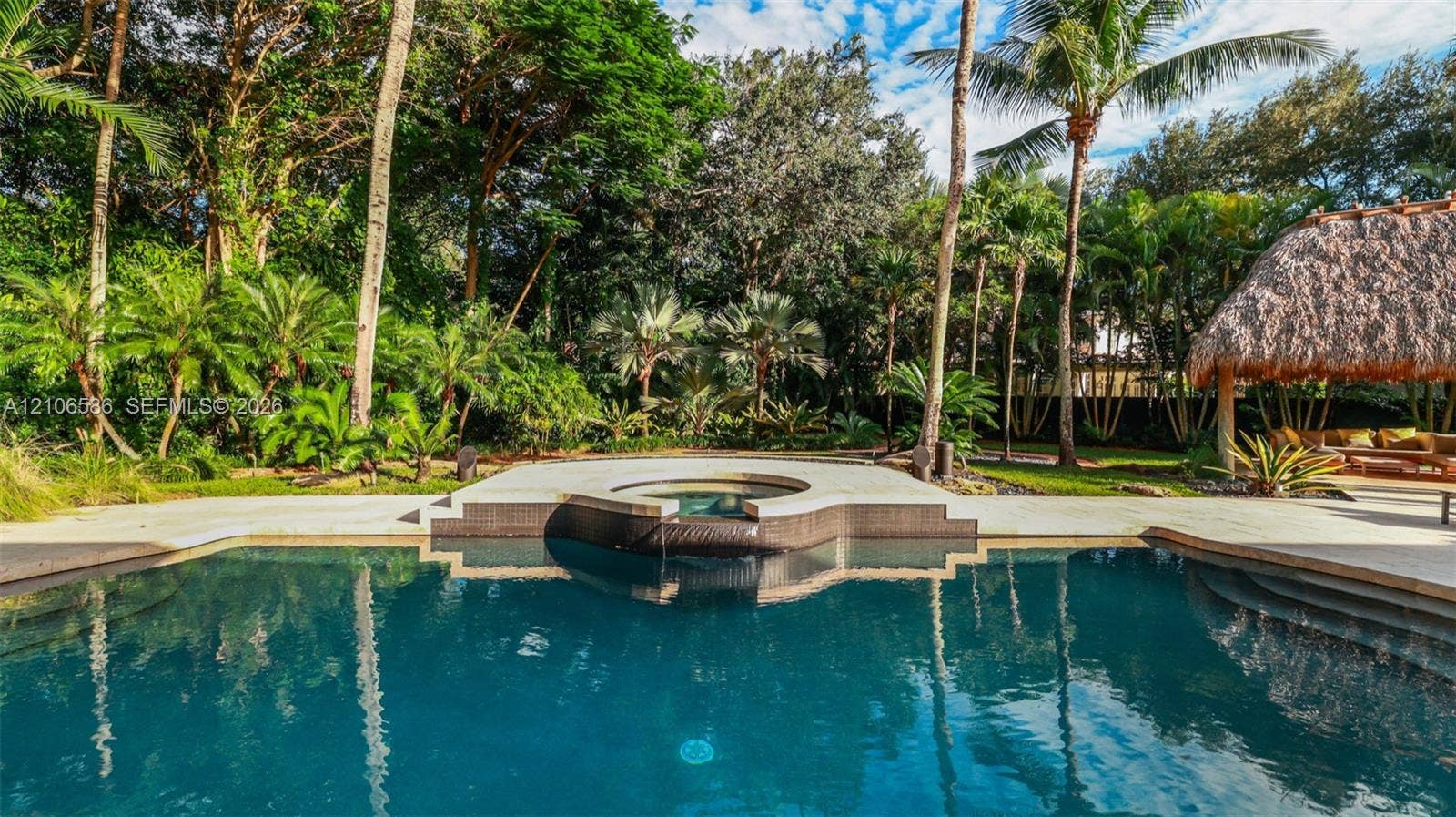 9325 SW 59th Place, Pinecrest, FL 33156 In-ground swimming pool featuring a tiered spillover spa, stone-tile surround, and a thatched-roof palapa set against a landscaped backdrop of mature palms and dense foliage