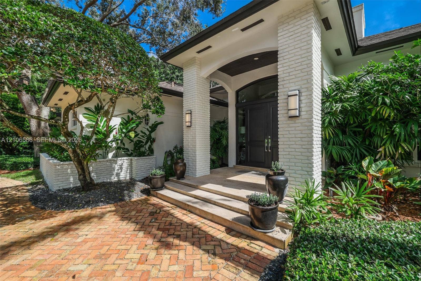 9325 SW 59th Place, Pinecrest, FL 33156 Covered entryway featuring white brick pillars, a multi-paneled dark wood front door with arched transom, tiered concrete steps, and a herringbone-patterned brick paver walkway