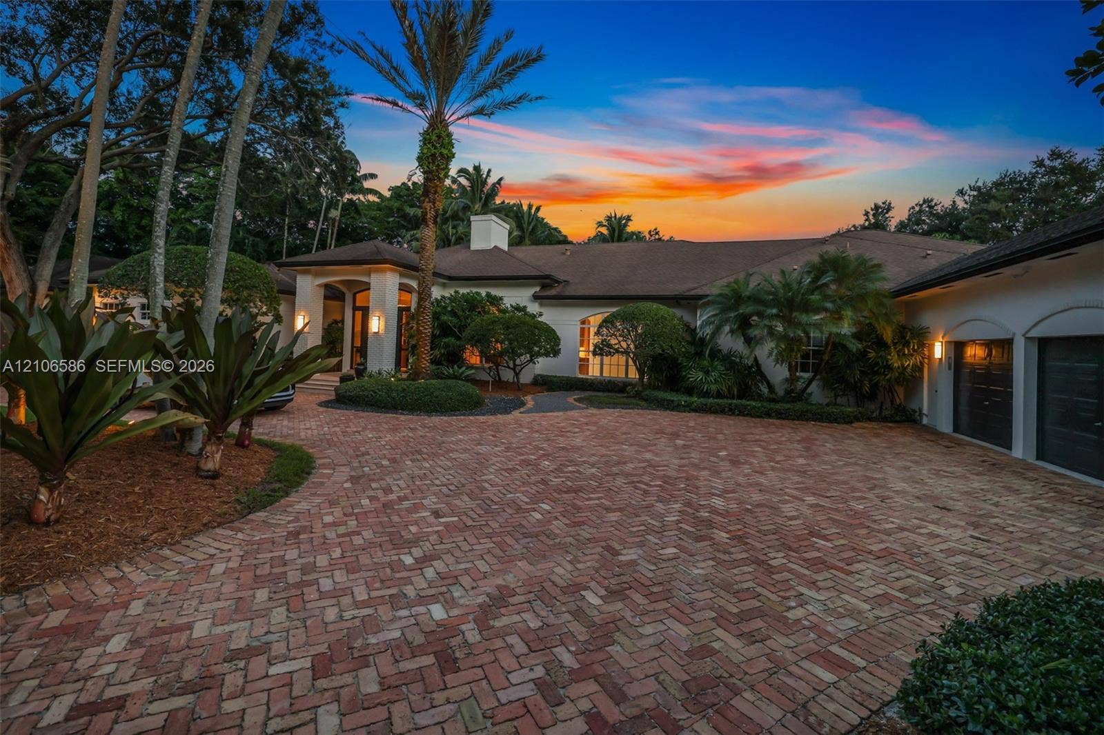 9325 SW 59th Place, Pinecrest, FL 33156 Stucco exterior featuring a herringbone-pattern brick paver driveway, covered entryway, multi-car garage, and mature landscaping