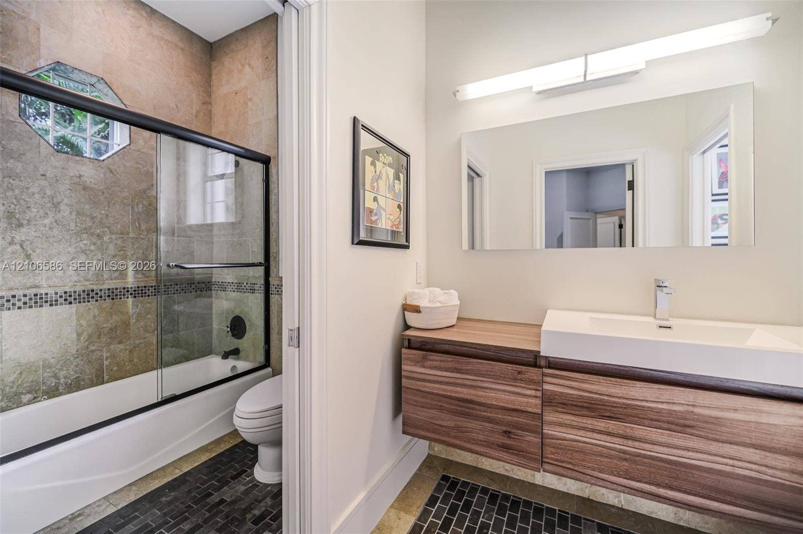 9325 SW 59th Place, Pinecrest, FL 33156 Floating wood-finish vanity with a rectangular integrated sink, wall-mounted rectangular mirror, shower enclosure featuring a glass sliding door, and stone-tiled shower surround