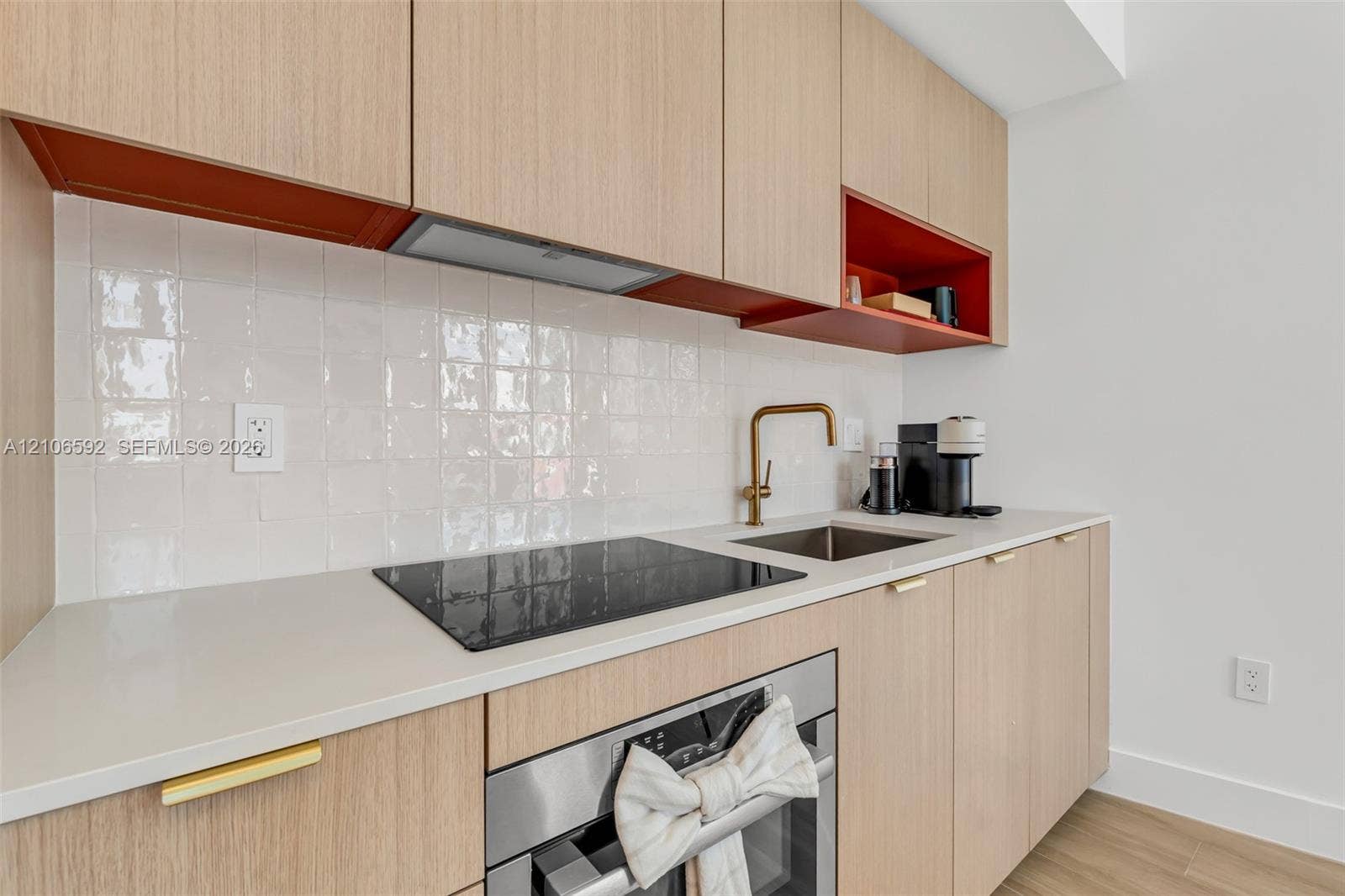 90 NE 32, #612, Miami, Miami, FL 33127 Flat-panel cabinetry with wood-finish veneer, white square-tile backsplash, light-toned solid surface countertop, built-in black induction cooktop, and brass-finish faucet