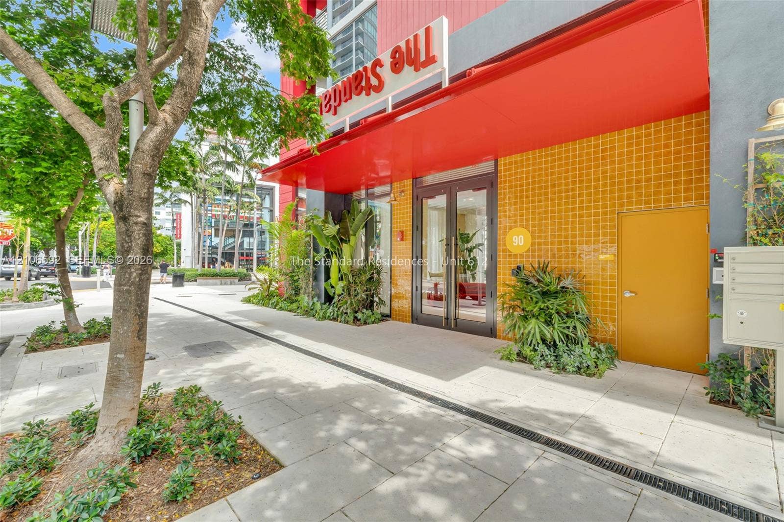 90 NE 32, #612, Miami, Miami, FL 33127 Building exterior featuring a vibrant yellow tiled facade, a prominent red metal awning, glass double-door entrance, and integrated landscape planting beds