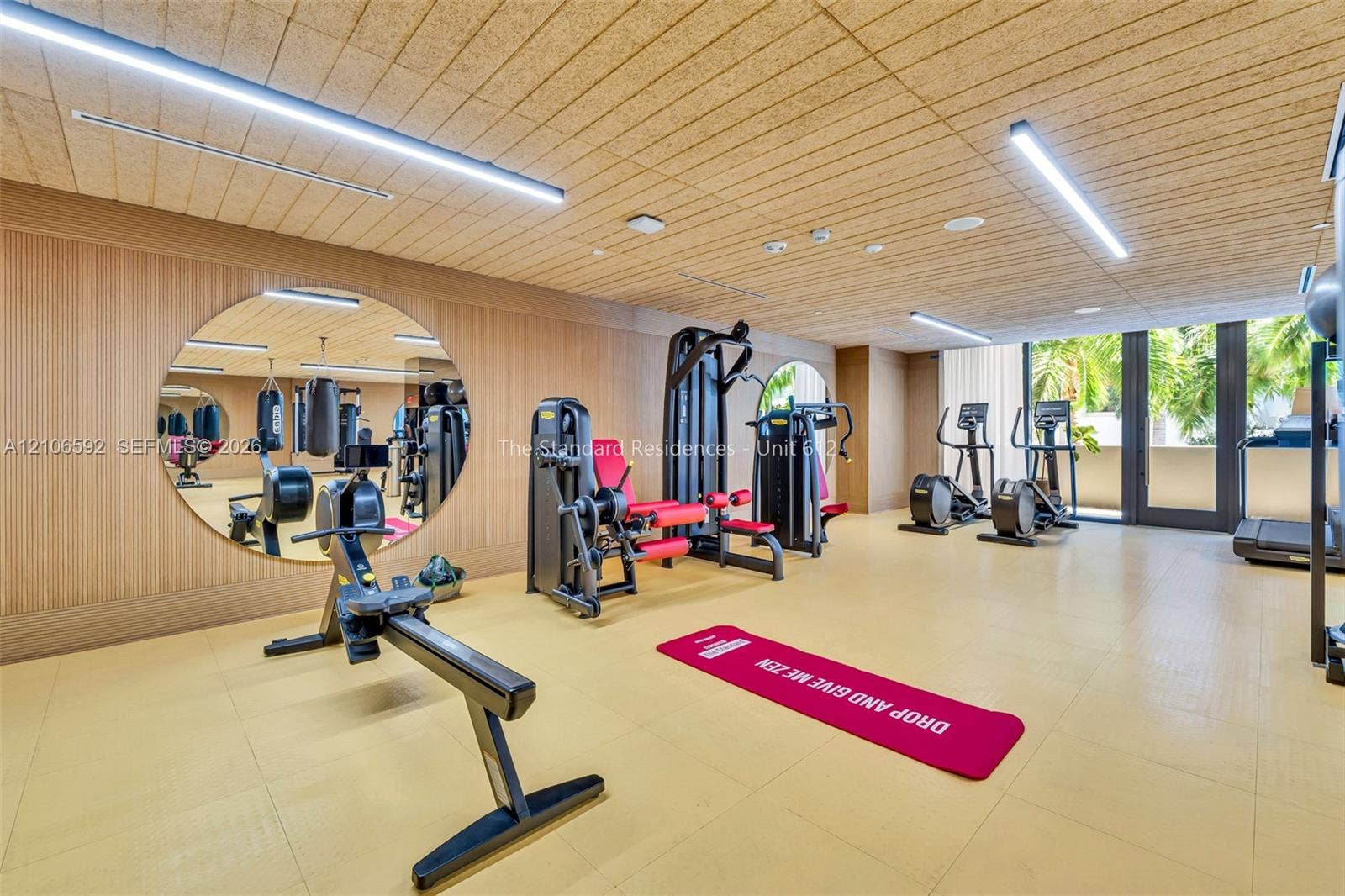 90 NE 32, #612, Miami, Miami, FL 33127 Fitness center featuring wood-paneled walls, a large circular wall-mounted mirror, linear LED lighting, and glass doors with exterior foliage