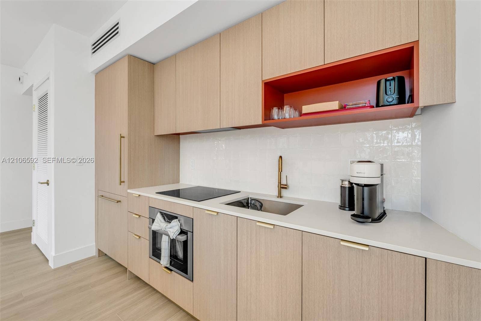 90 NE 32, #612, Miami, Miami, FL 33127 Flat-panel wood-finish cabinetry, white solid-surface countertop, integrated electric cooktop, stainless steel under-mount sink with brass-toned faucet, and gloss-finish wall tiling