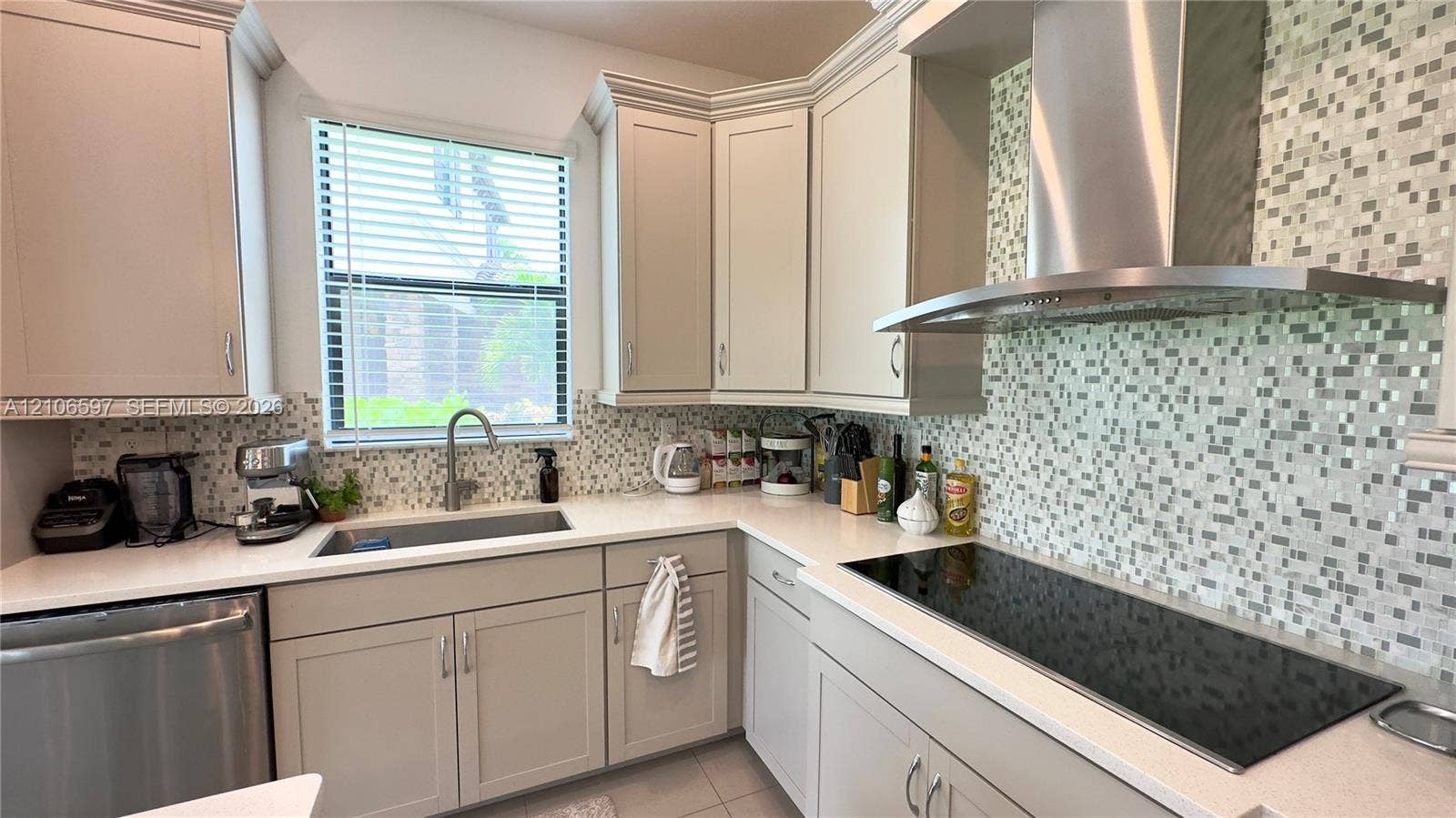 Kitchen featuring neutral cabinetry, white stone-finish countertops, multi-tonal mosaic tile backsplash, stainless steel dishwasher, and stainless steel wall-mounted range hood