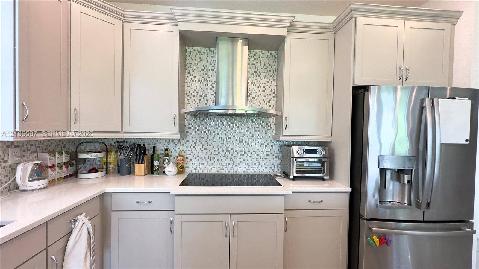 Kitchen featuring neutral-toned cabinetry, solid surface countertops, a mosaic tile backsplash, a stainless steel chimney-style range hood, and a built-in stainless steel refrigerator