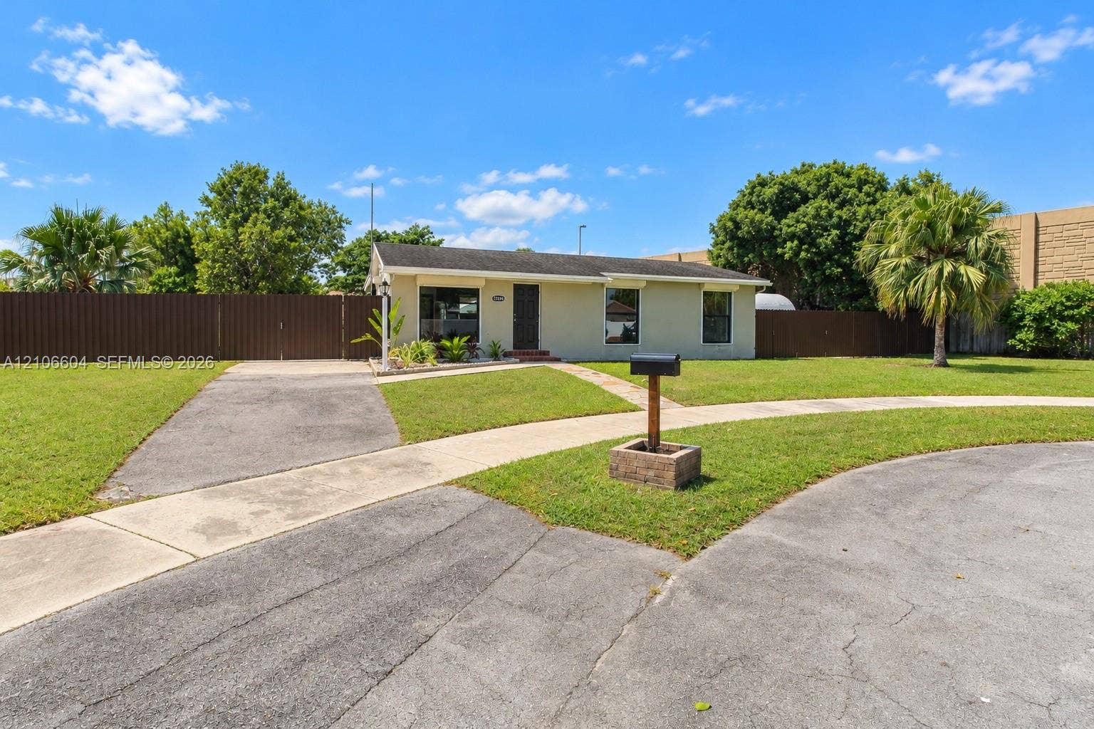 Single-story exterior featuring a light-toned stucco facade, asphalt shingle roof, expansive front lawn, paved driveway, and privacy fencing