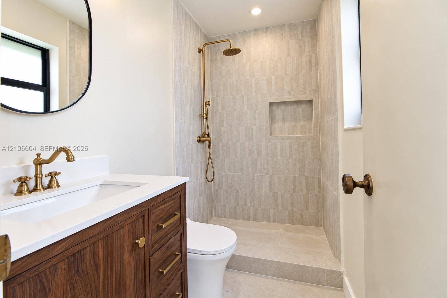 Wood-finish vanity with white countertop, brass-toned fixtures, walk-in shower with vertical tile surround and built-in niche, and brass hardware accents