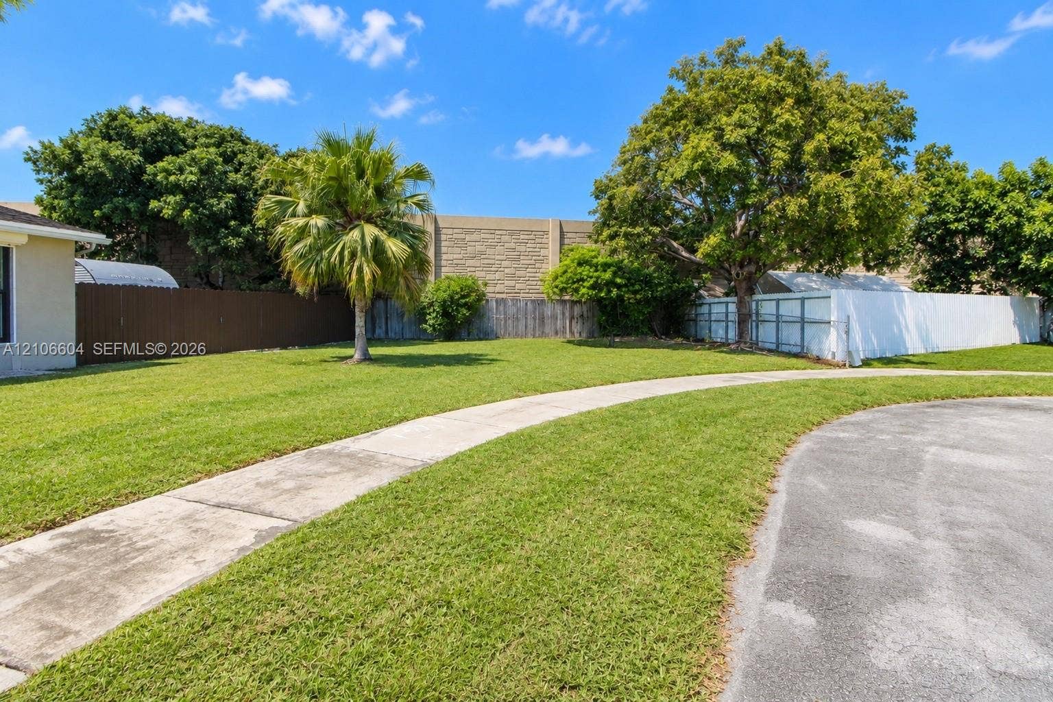 Expansive lawn featuring mature trees, palm landscaping, concrete walkway, and perimeter privacy fencing