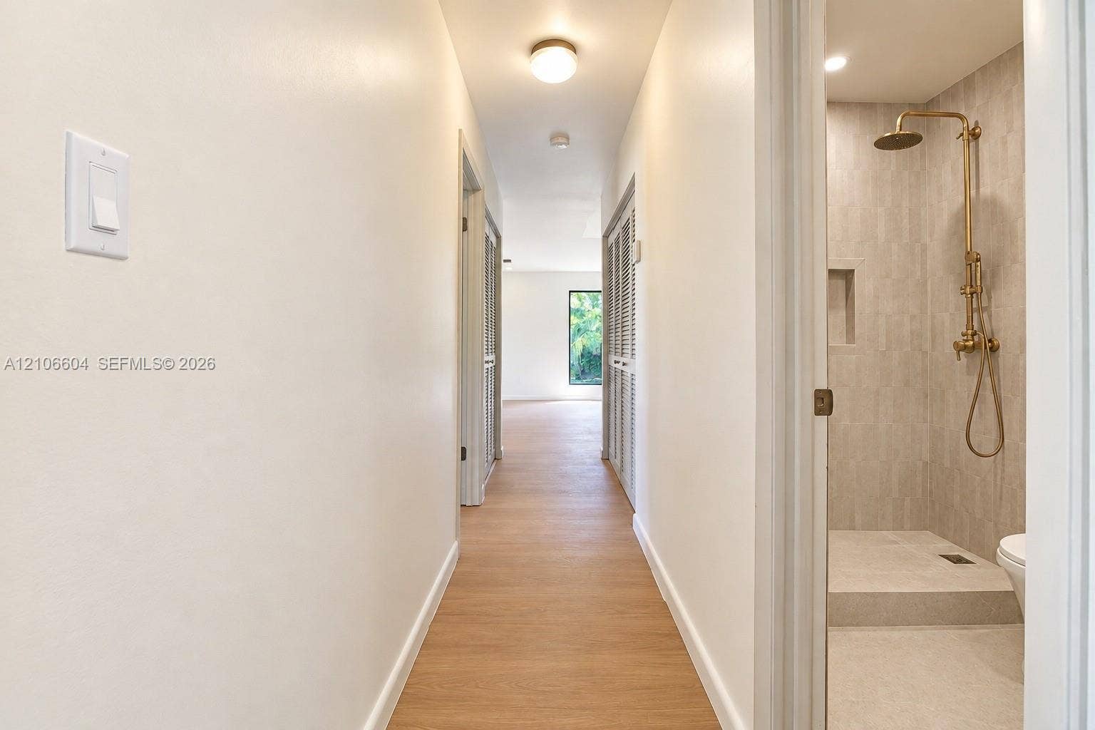 Hallway featuring wood-finish flooring and flush-mount lighting, with a bathroom entry showcasing a tiled walk-in shower, built-in wall niche, and brass-finish fixtures