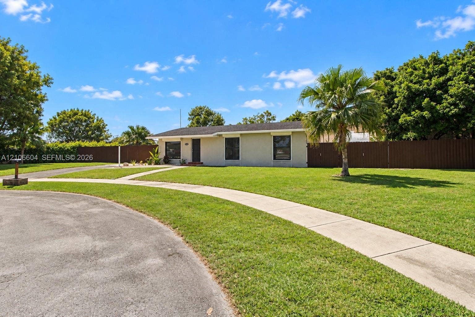Single-story residence featuring a low-slope roof, light-colored exterior finish, concrete walkway, and expansive grassy front lawn enclosed by perimeter fencing